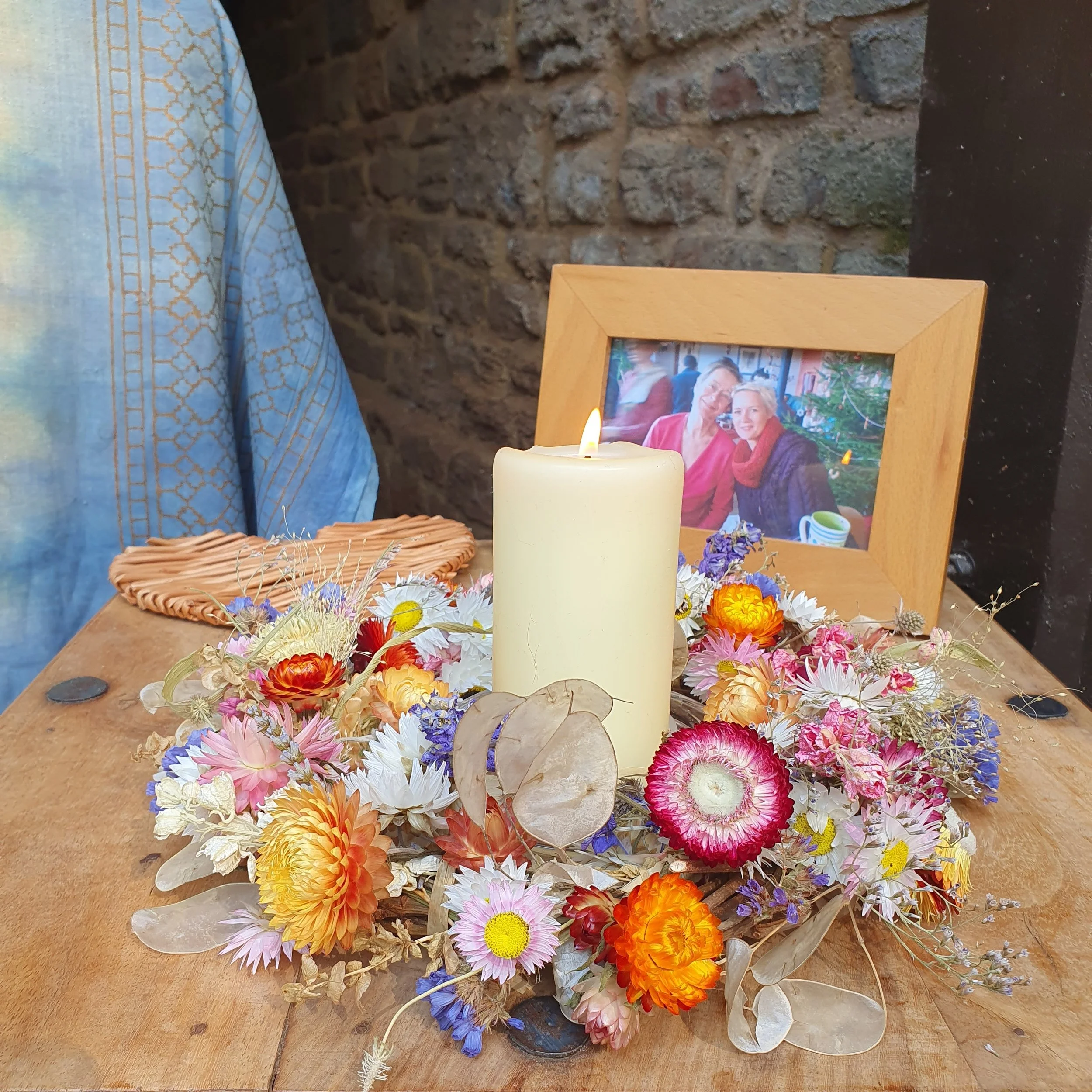A memorial display with a photo of two women, candles, and an array of colorful flowers on a wooden surface against a brick wall.