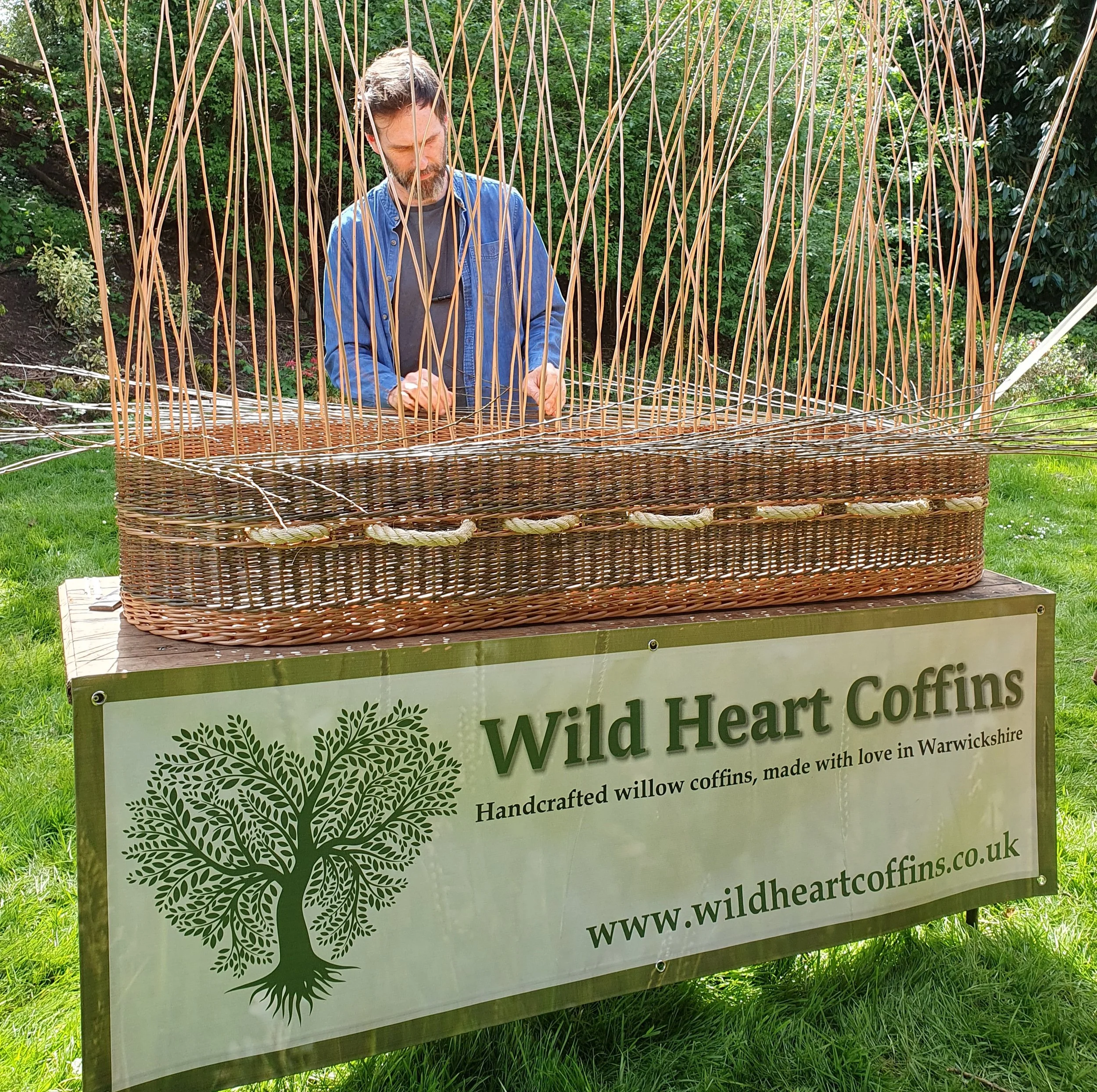 A man with a beard and dark hair, wearing a denim shirt and grey t-shirt, standing behind a woven willow coffin with a sign for Wild Heart Coffins. The setting is outdoors with green trees and grass.