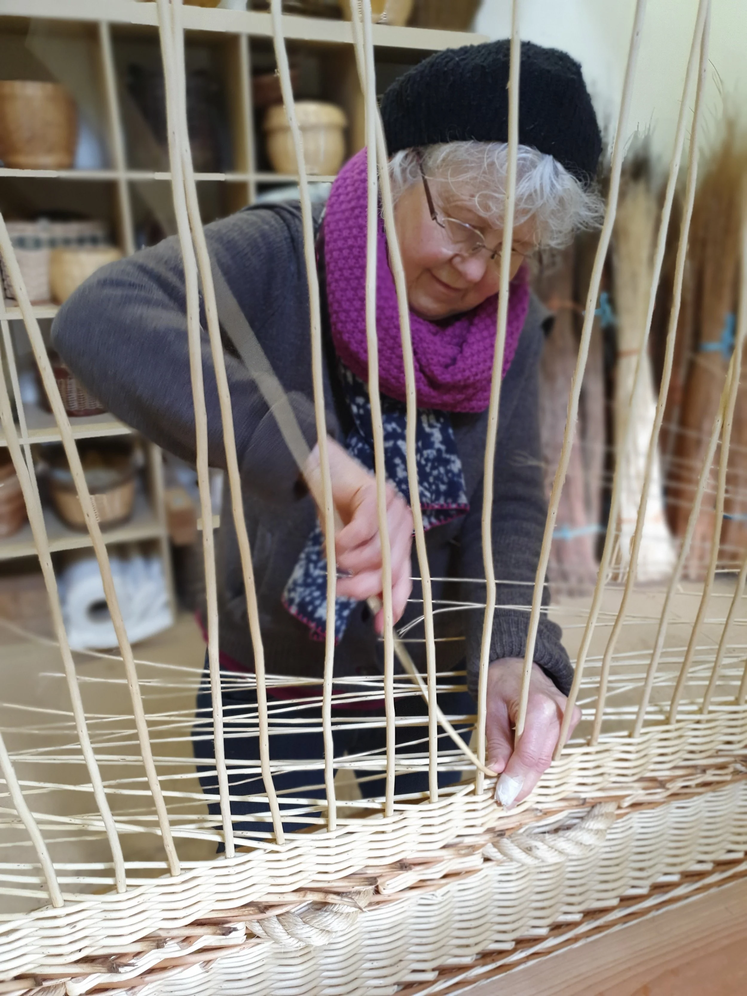 An elderly woman with gray hair and glasses, wearing a black hat, pink scarf, and gray sweater, is working with her hands inside a large woven basket or cage, possibly involved in crafting or basket-making.