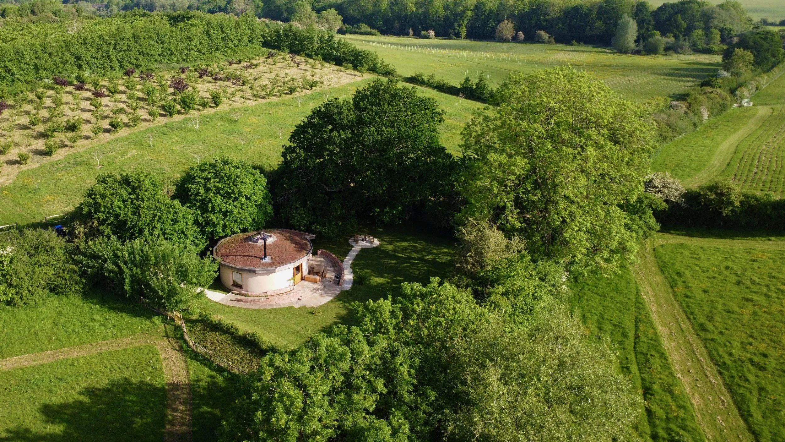 Aerial view of a lush green landscape with various trees, grassy fields, and a small round building with a circular patio surrounded by greenery.