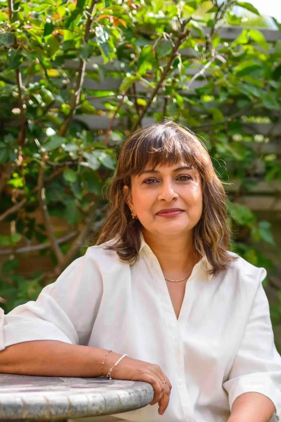 A woman with shoulder-length brown hair wearing a white blouse, jewelry, and makeup, sitting outdoors at a table in front of green leafy bushes under bright sunlight.