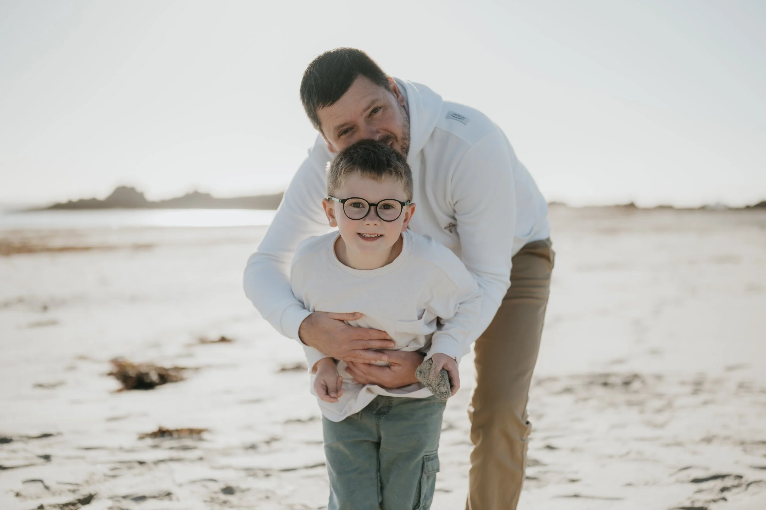 Un homme et un garçon jouent sur la plage. L'homme, agenouillé, tient le garçon par la taille. Le garçon porte des lunettes rondes et un t-shirt blanc. Le soleil brille et le paysage est légèrement flou en arrière-plan.