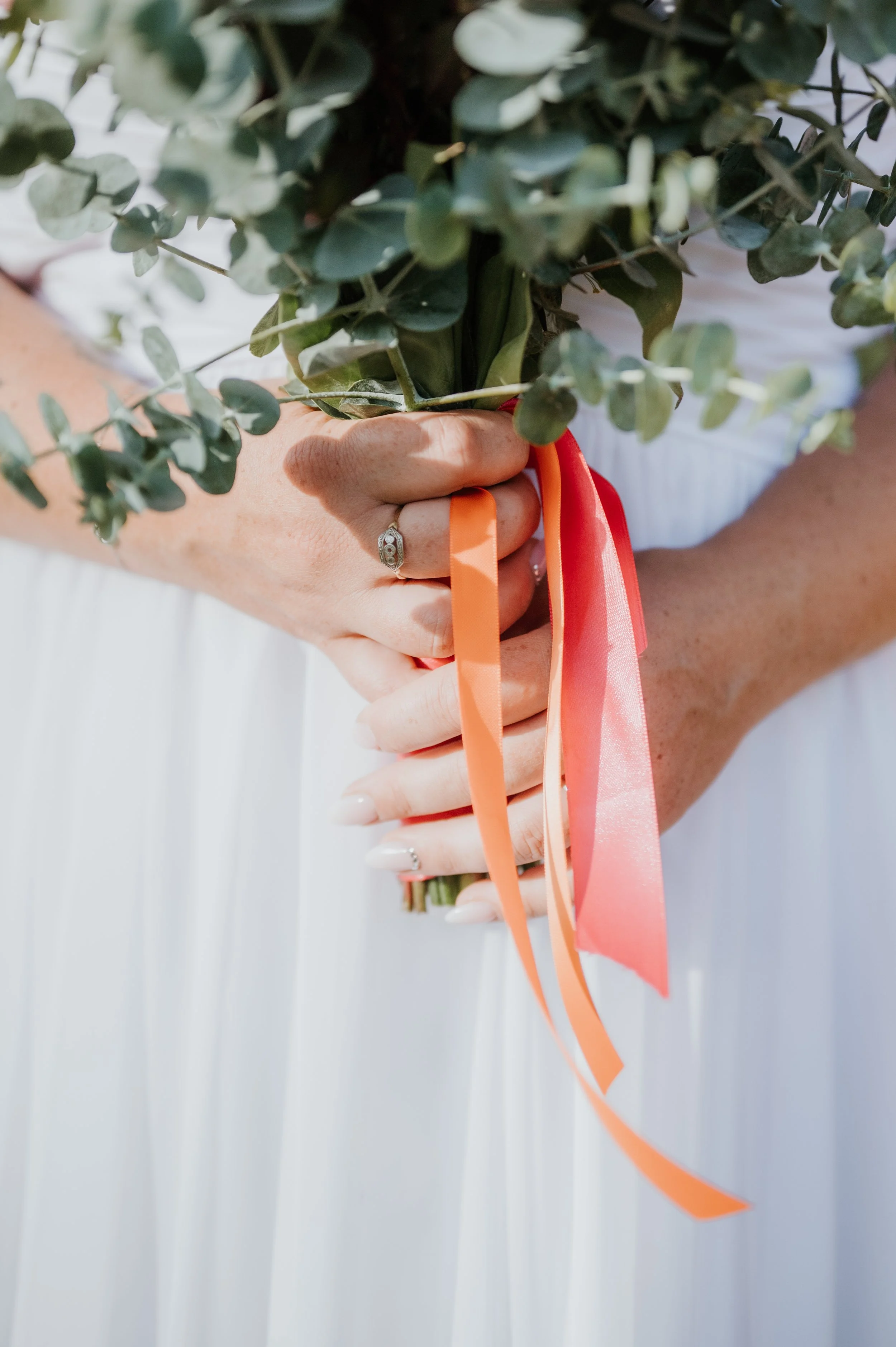 Une main tenant un bouquet de fleurs vertes avec des rubans orange et rose, portant une bague en diamant, contre un fond blanc.