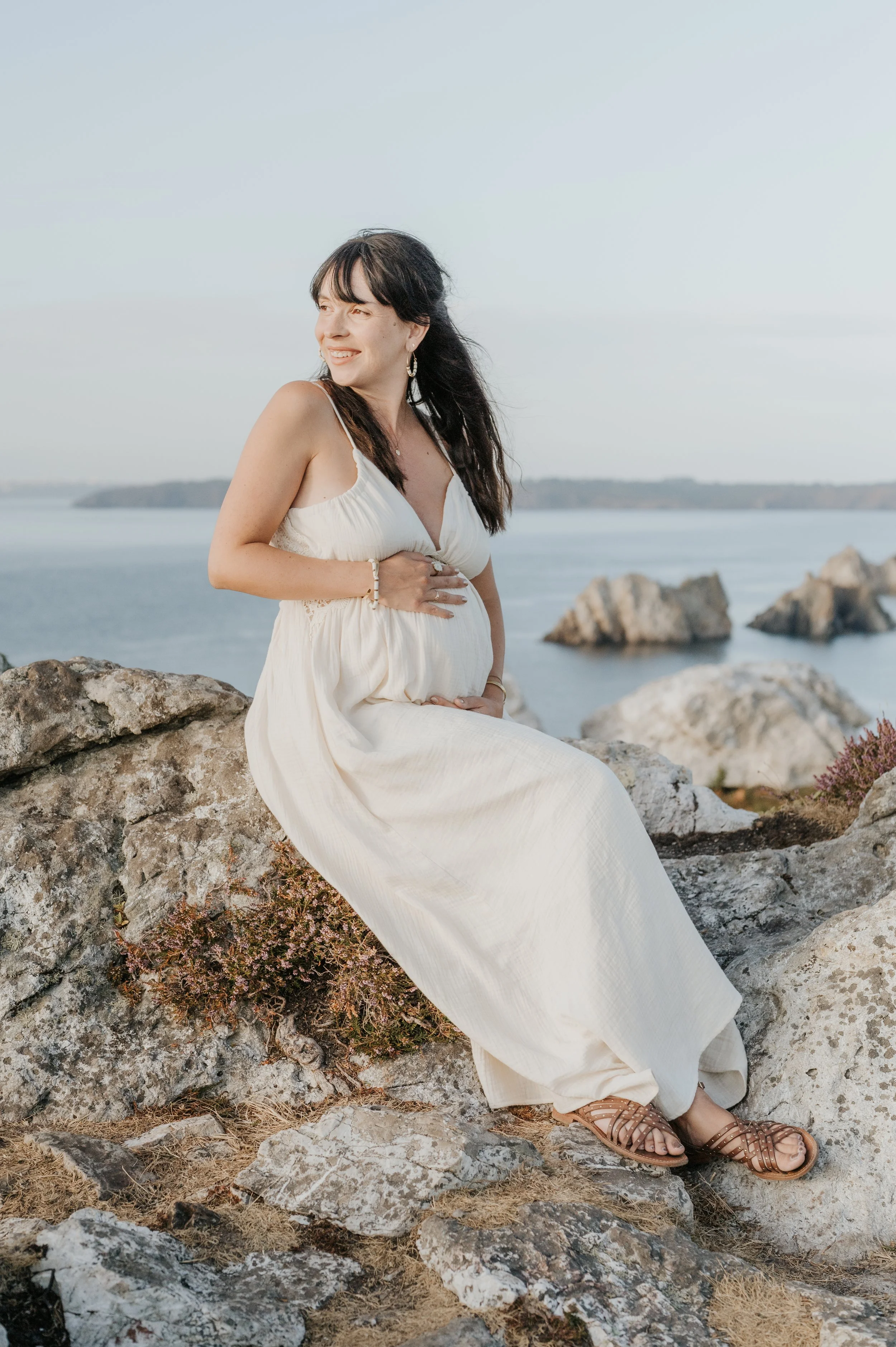 Femme enceinte assise sur des rochers en bord de mer, portant une robe blanche, en regardant vers la gauche, avec un ciel clair en fond.