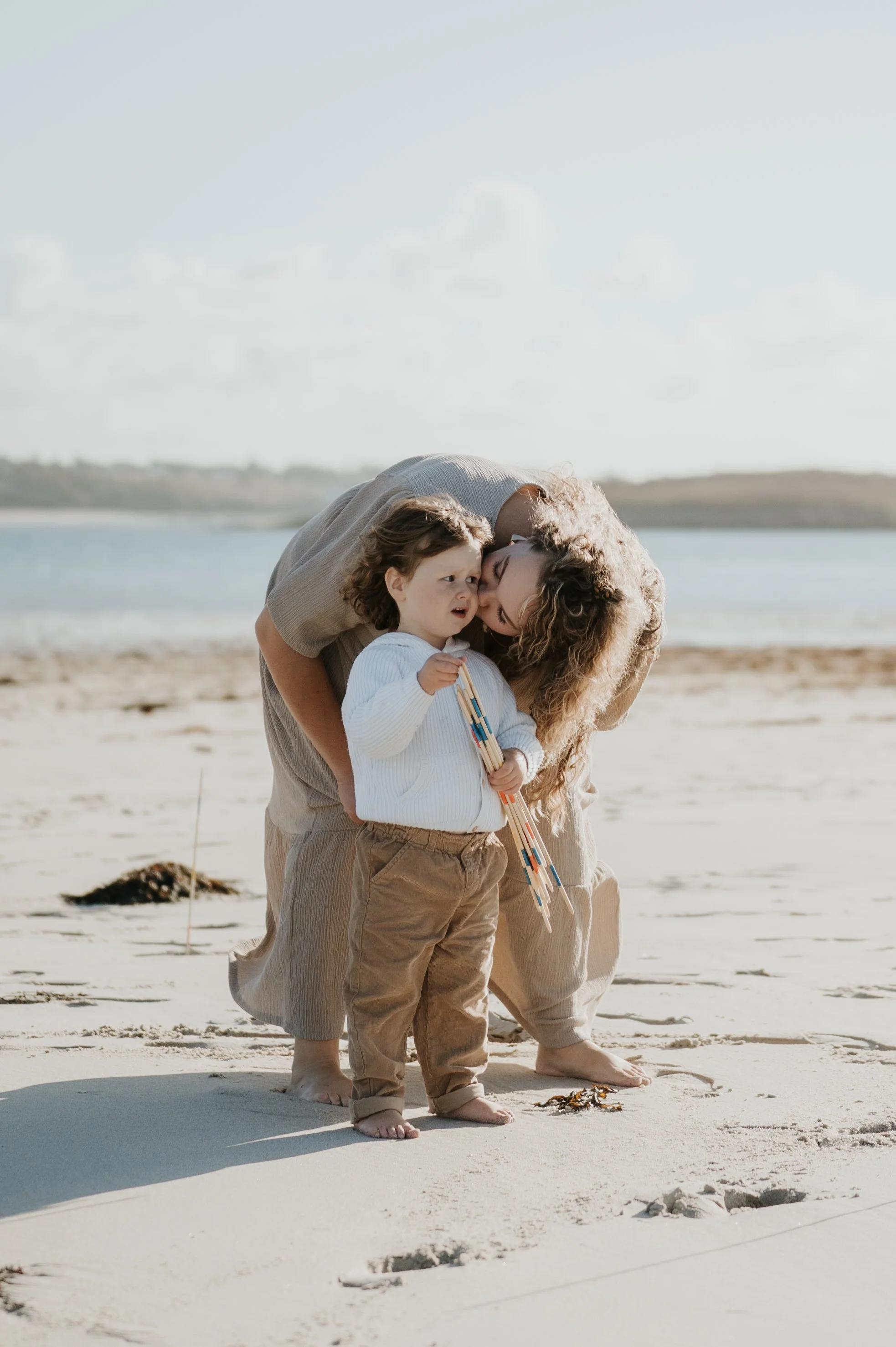 Une famille de trois personnes, deux adultes et un enfant, se tient sur la plage, partageant un moment d'affection et de tendresse, avec la mer en arrière-plan.