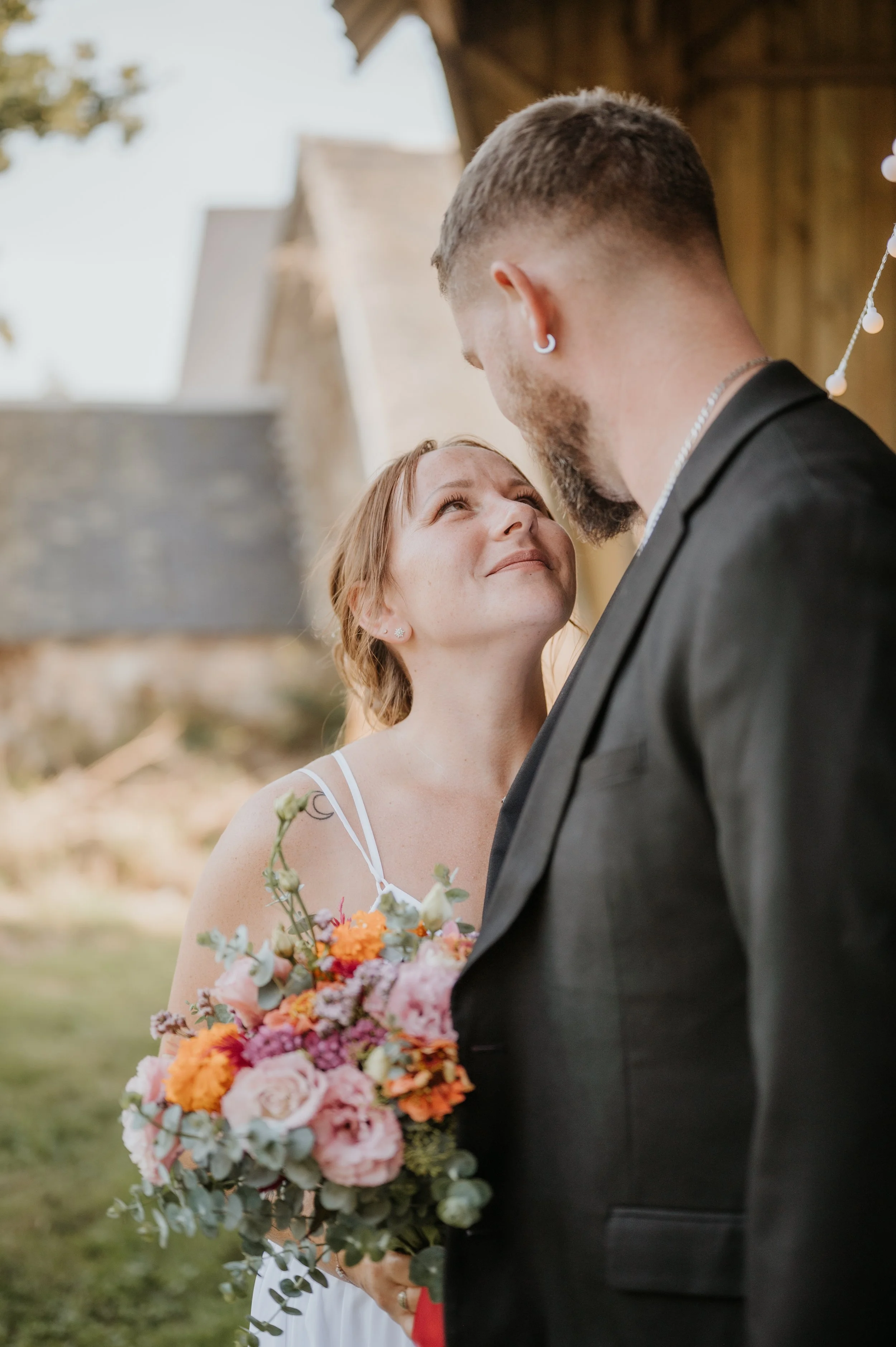 Un couple lors de leur mariage, la femme regarde l'homme avec affection, la mariée tient un bouquet de fleurs colorées, la scène se déroule en plein air avec une structure en bois en arrière-plan.