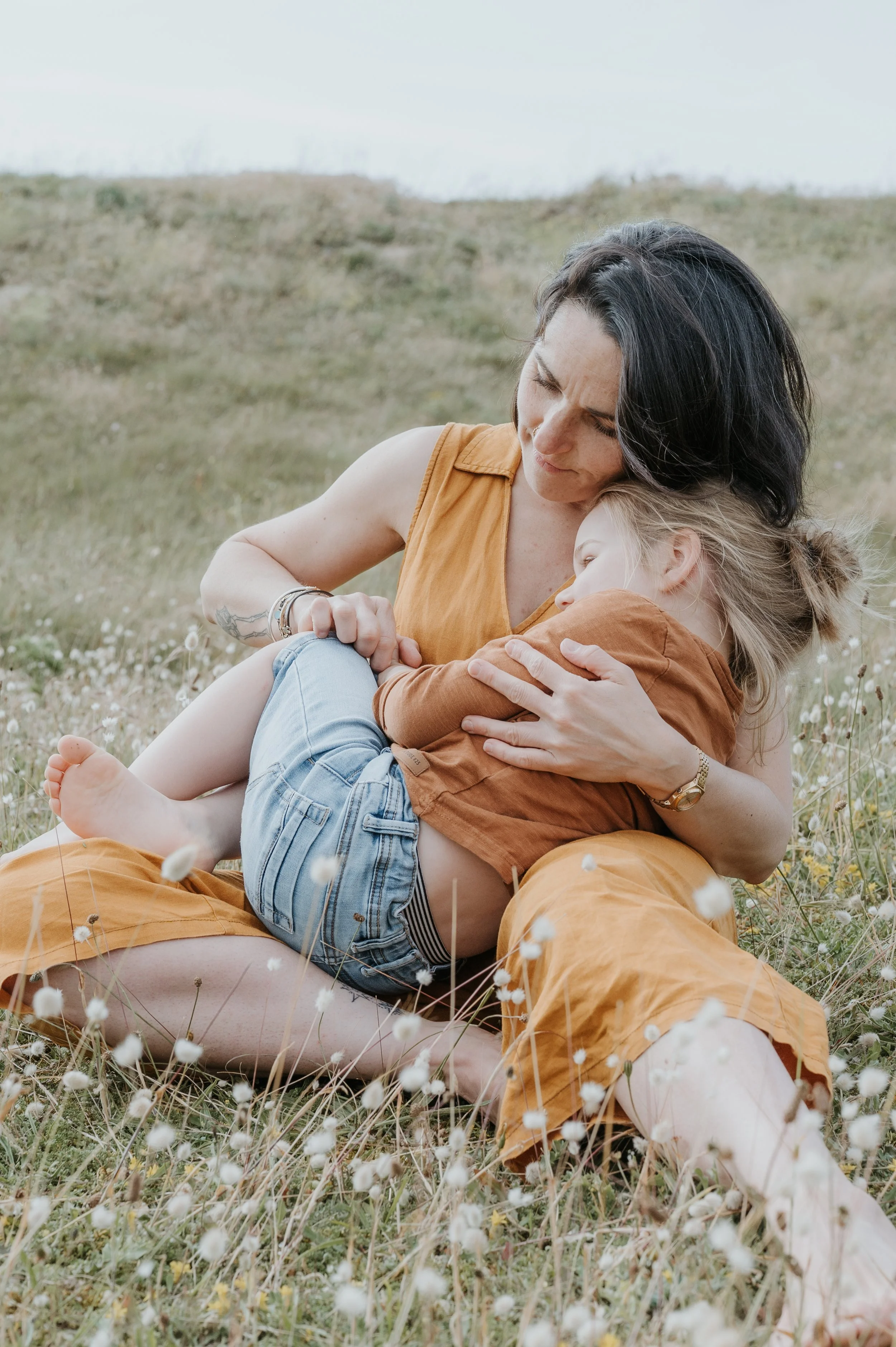Une femme et son fils partagent un moment tendre en plein air dans un champ de fleurs sauvages, assises sur l'herbe, avec un paysage de collines en arrière-plan.