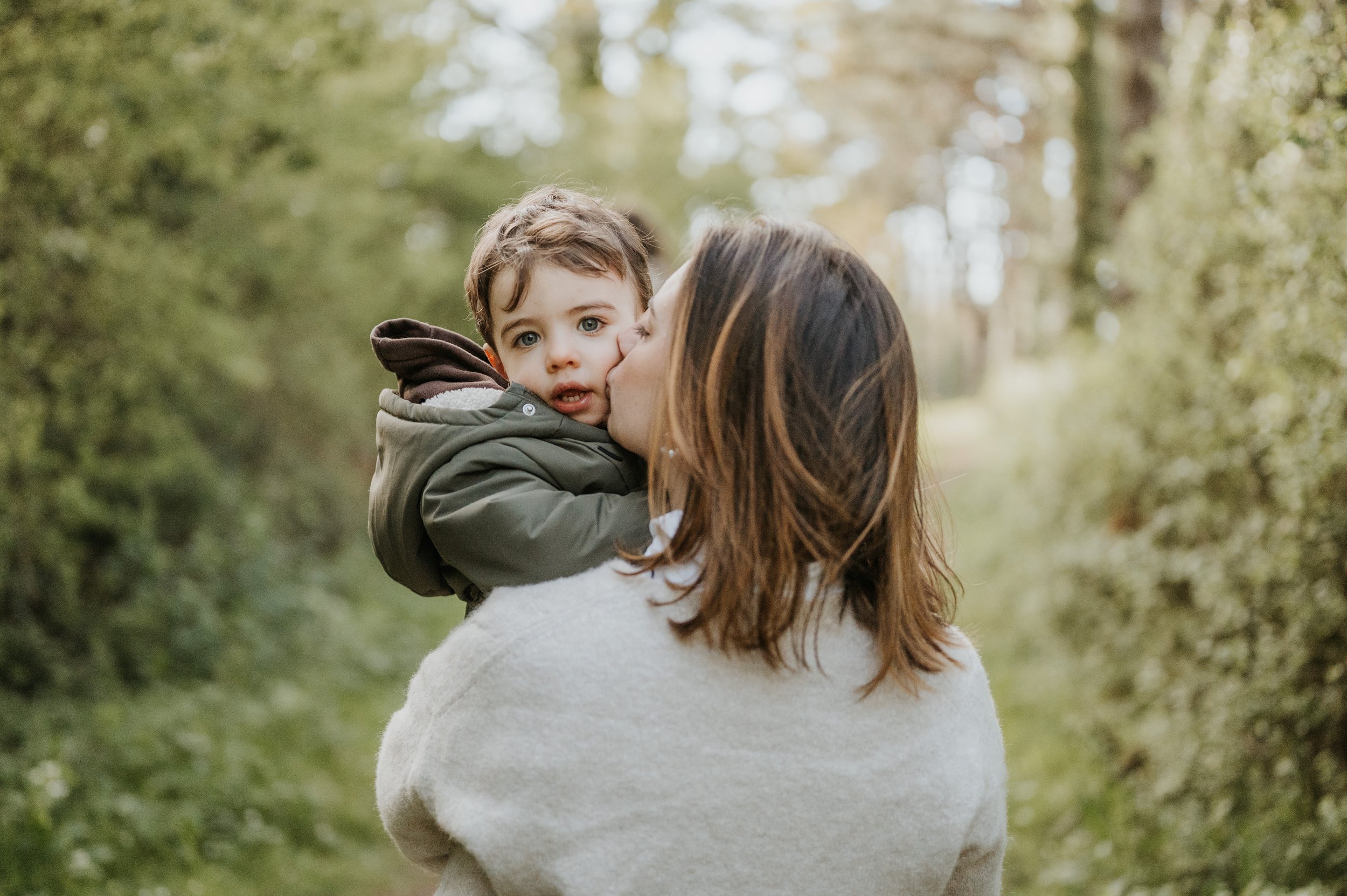 Une femme donne un câlin à un jeune garçon dans un environnement naturel, probablement une forêt, avec des arbres et des feuilles vertes en arrière-plan.
