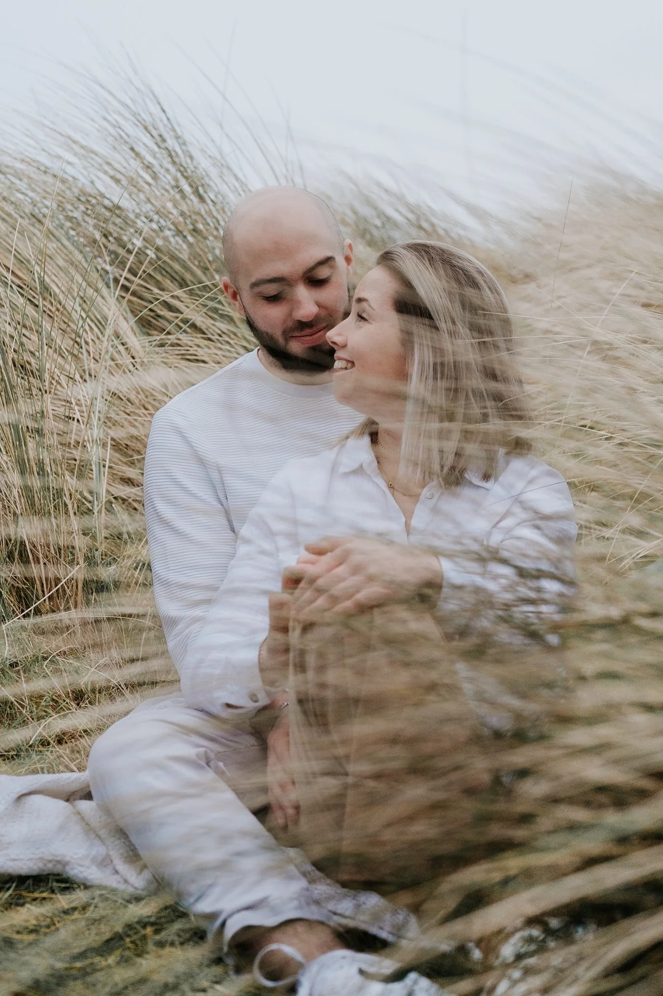 Un couple souriant assis dans un champ de hautes herbes, se regardant tendrement. La femme porte une chemise blanche et des lunettes, l'homme porte un t-shirt rayé blanc et beige. La scène est en plein air, dans un environnement naturel et paisible.