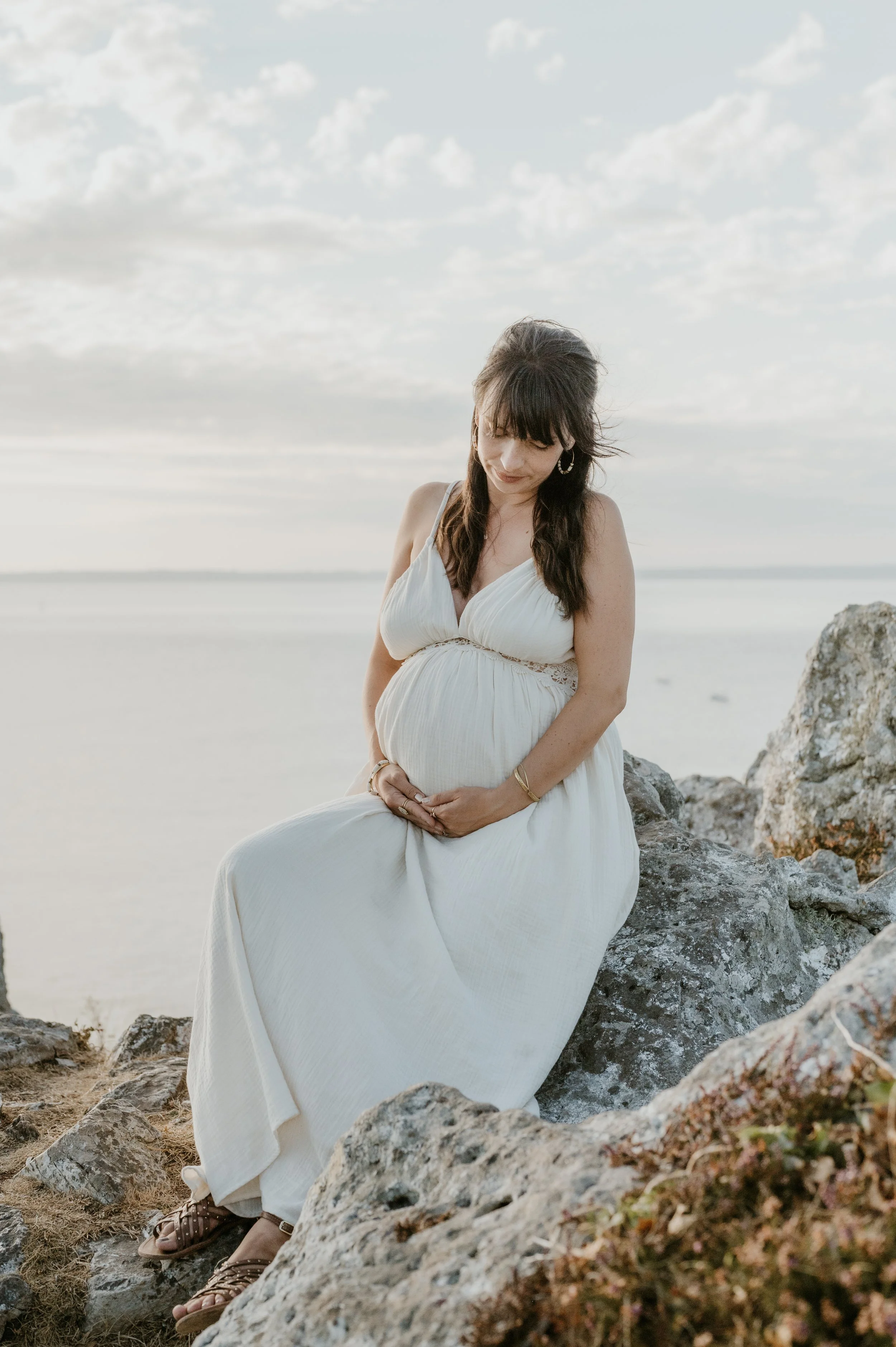 Une femme enceinte assise sur des rochers au bord de la mer par une journée ensoleillée, portant une robe blanche et regardant doucement son ventre. Le ciel est partiellement nuageux.