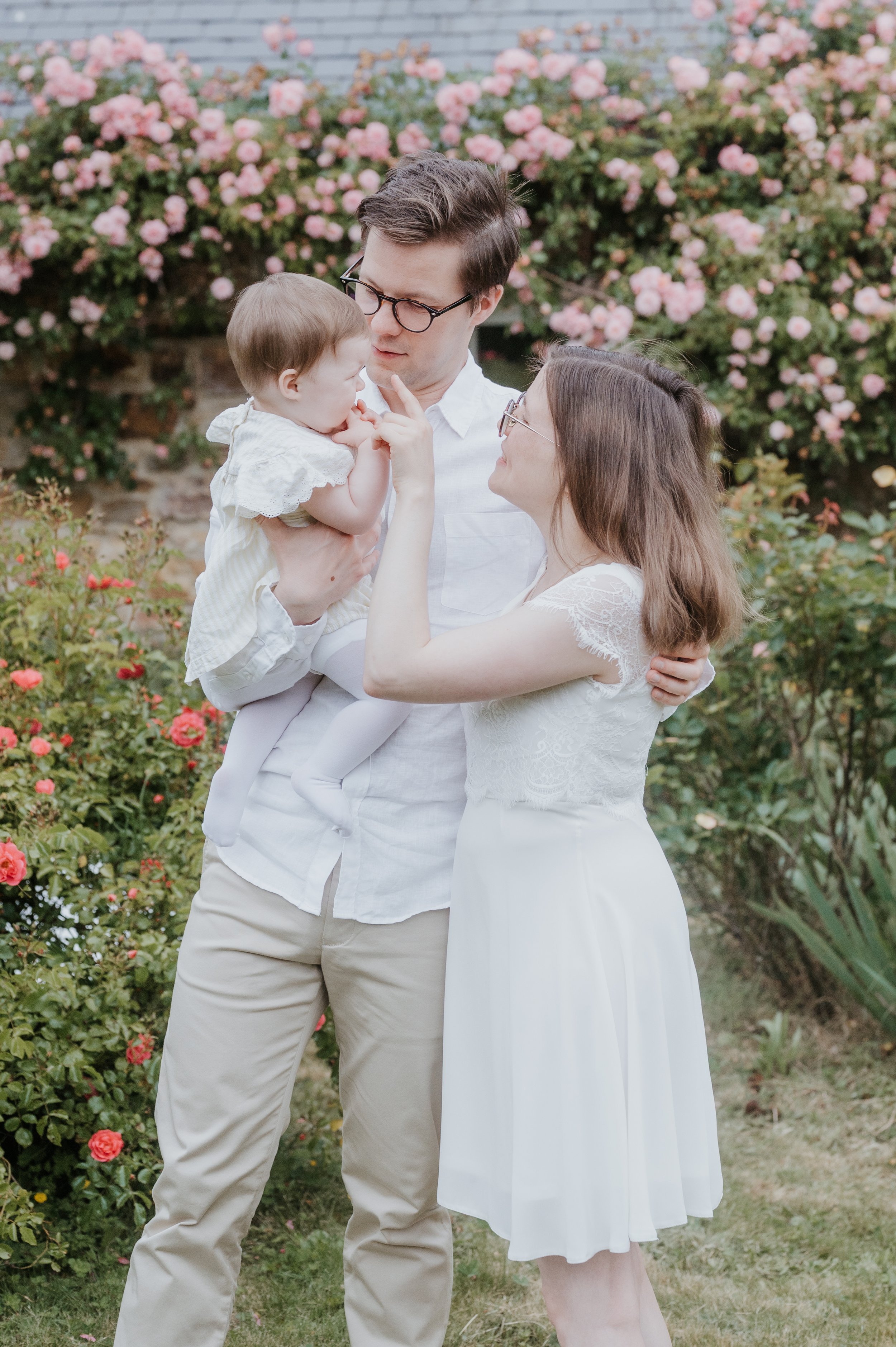 Une famille lors d'une séance photo en plein air devant un buisson de roses en fleurs. Un homme portant une chemise blanche et des pantalons beiges tient une jeune enfant blanche habillée d'une robe blanche. Une femme portant une robe blanche avec de