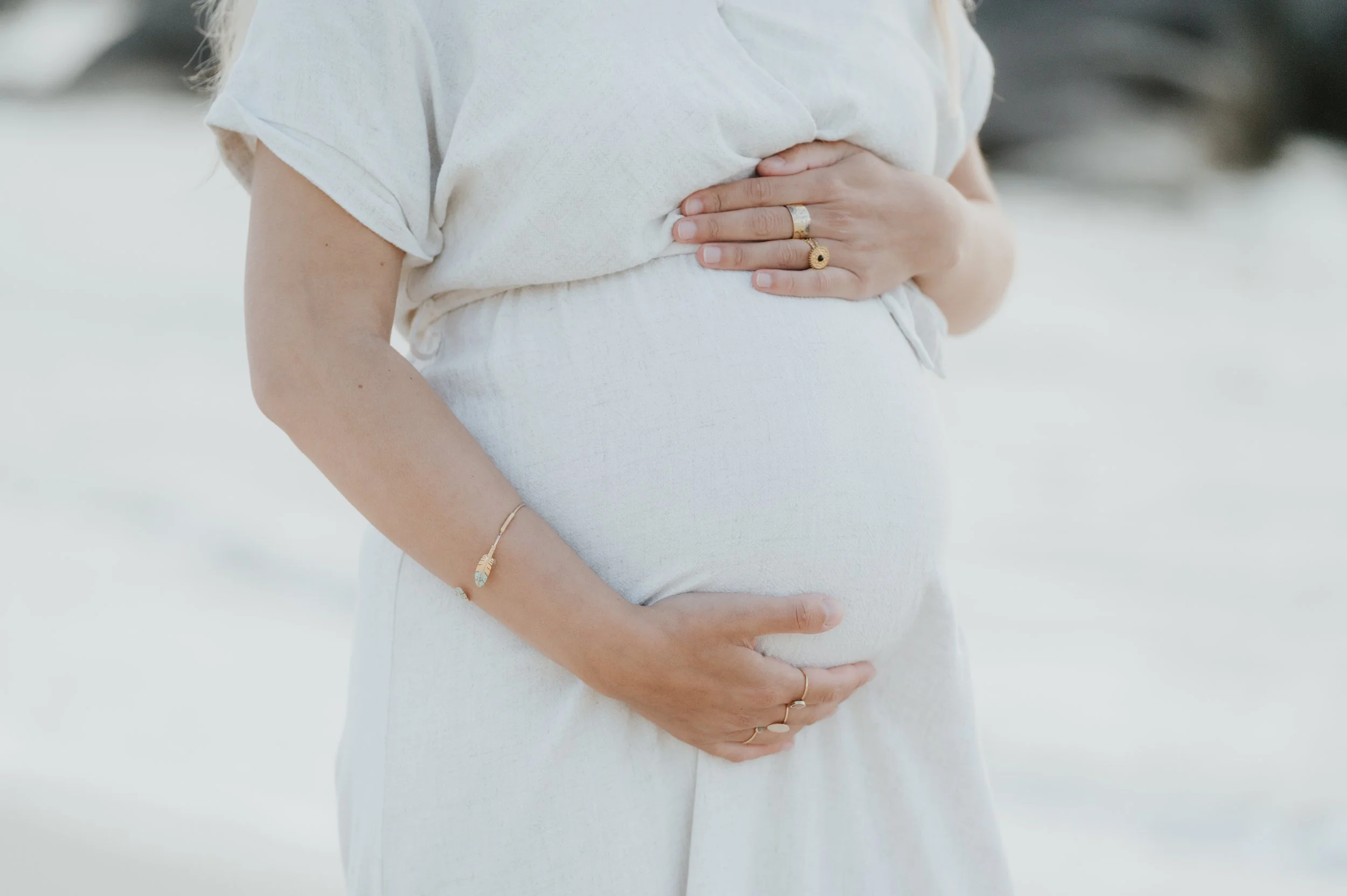 Une femme enceinte tient son ventre avec les mains, porte des bijoux en or et porte des vêtements blancs, dans un cadre naturel lumineux.