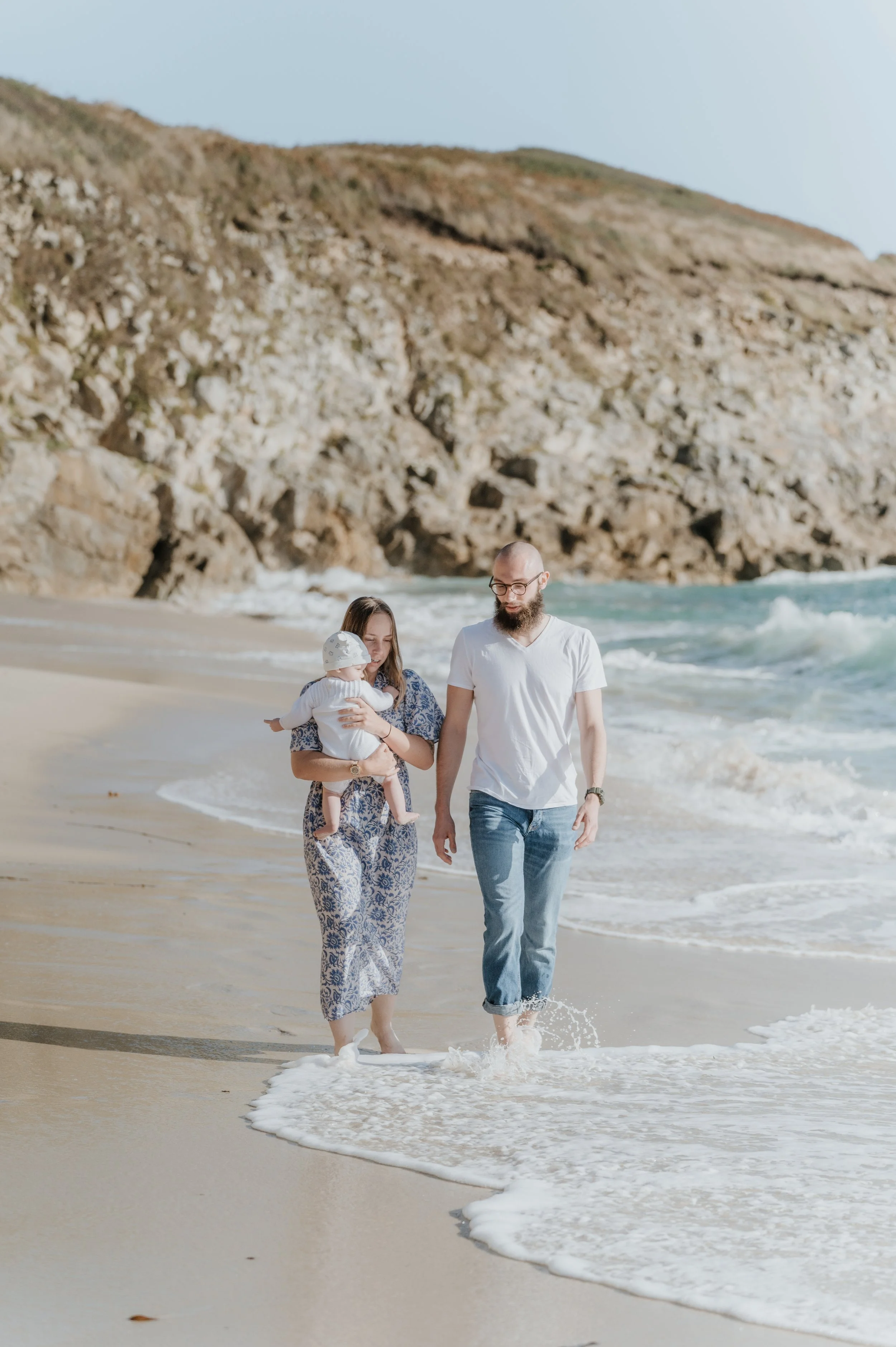 Un couple marche sur la plage avec un bébé. La femme tient le bébé, et tous deux sont habillés en vêtements légers. La mer et une falaise rocheuse sont visibles à l'arrière-plan, sous un ciel clair.