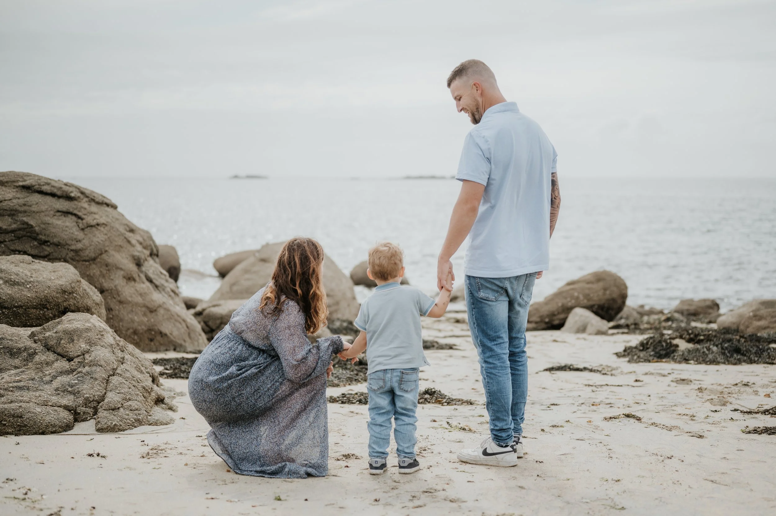 Une famille à la plage, un homme tenant la main d'un garçon, une femme à genoux, tous regardant le garçon, rochers en arrière-plan, ciel nuageux.