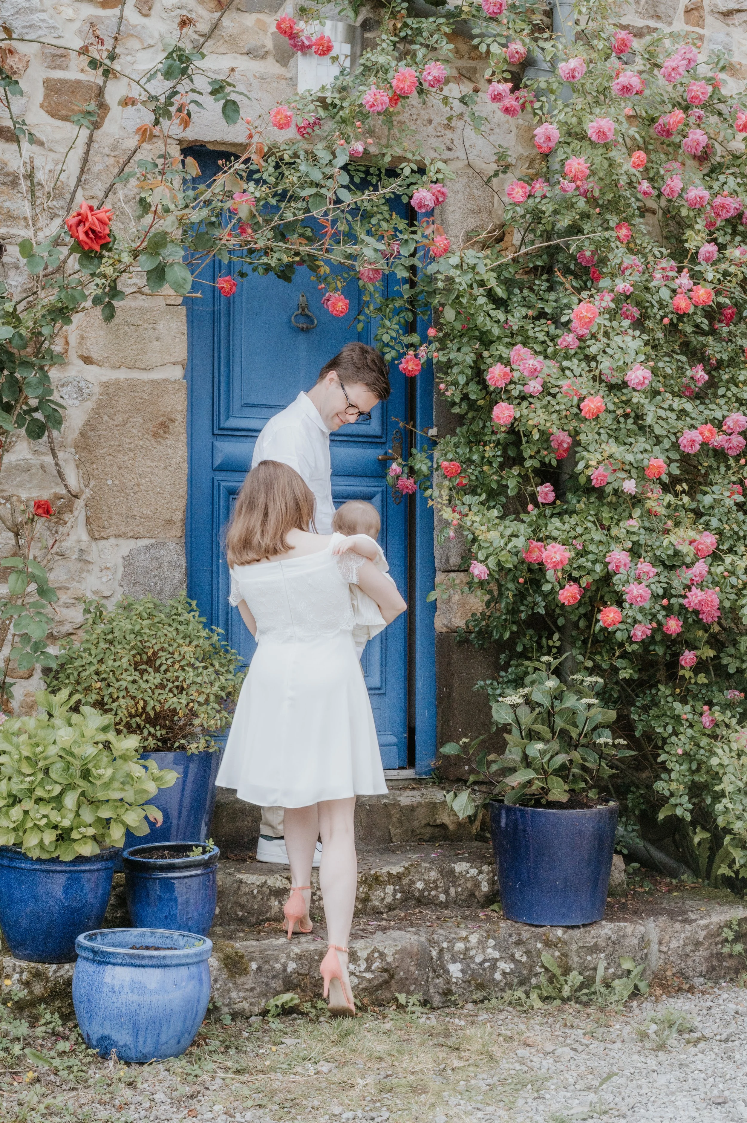 Un homme, une femme et un bébé en train de se tenir dans une cour pavée près d'une porte bleue entourée de roses roses dans des jardinières en céramique bleues.