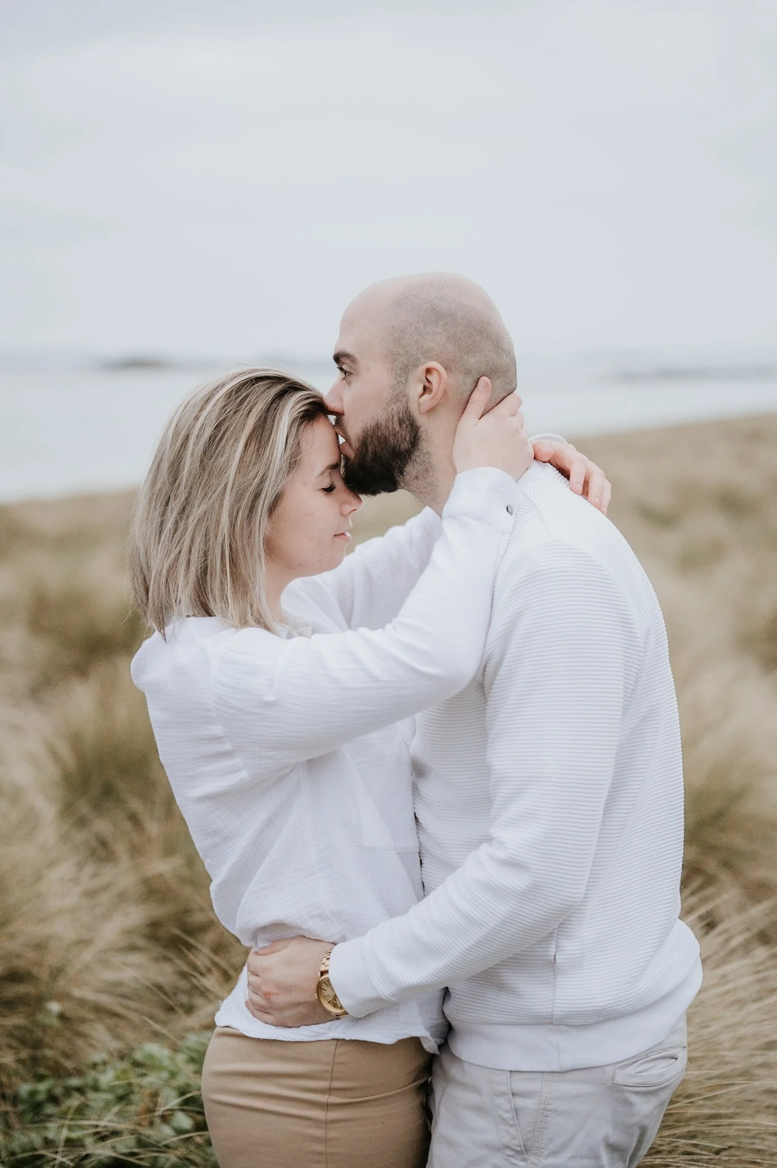 Un couple s'embrasse tendrement sur une plage, en arrière-plan un ciel gris et la mer calme.