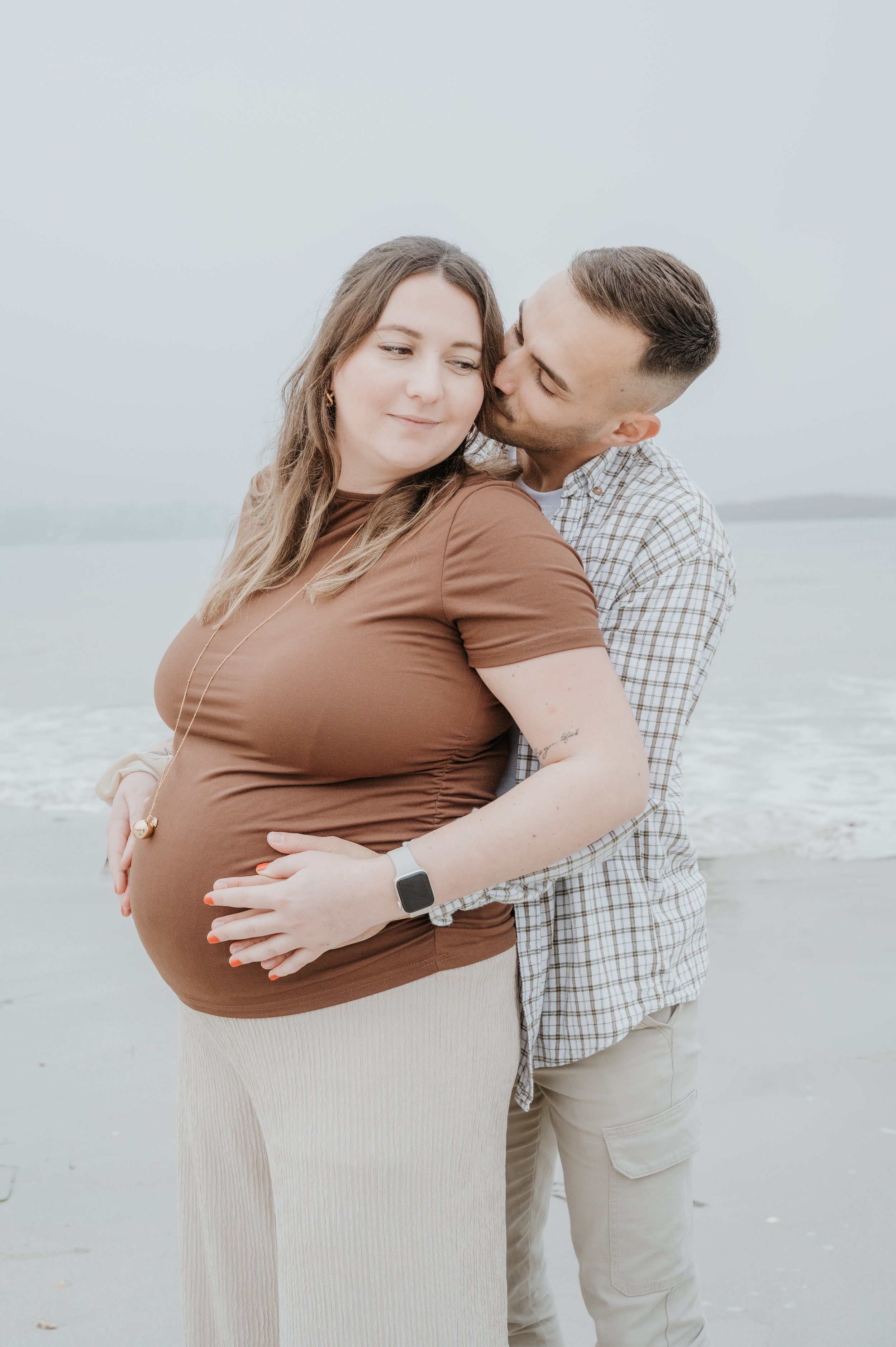 Un couple lors d'une séance photo sur la plage, la femme enceinte est en voiture, le homme l'entoure et l'embrasse tendrement, tous deux souriants.