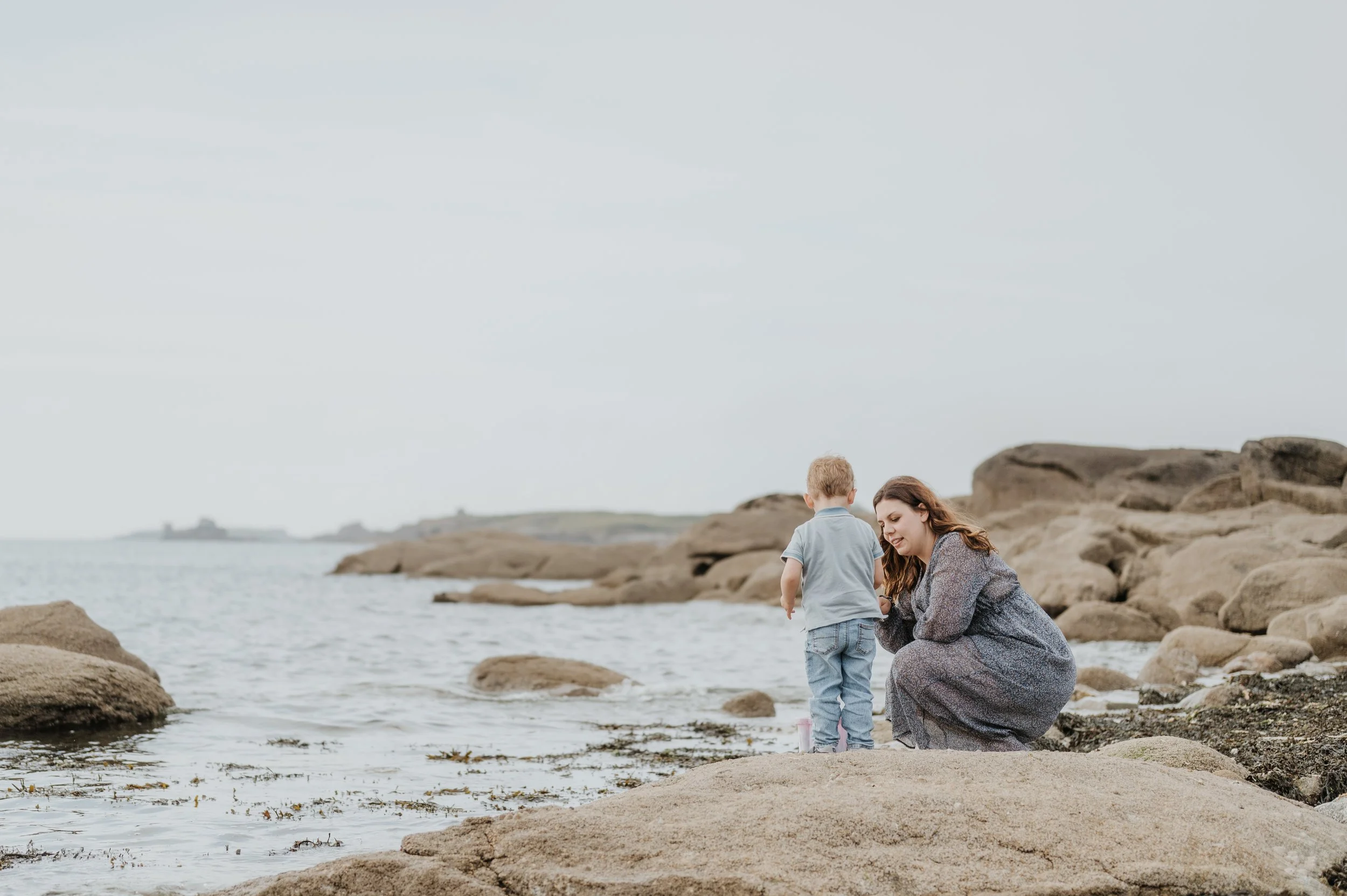 Une femme et un jeune garçon au bord de la mer, assis sur des rochers, en train de jouer dans le sable près de l'eau, avec un paysage côtier en arrière-plan.