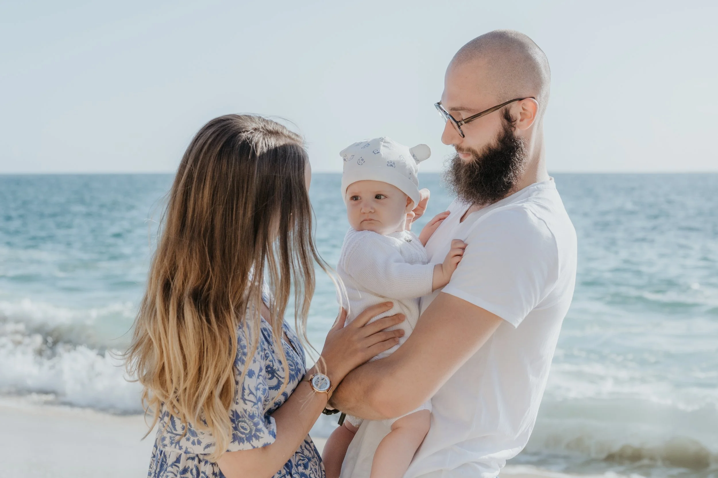 Un couple et un bébé regardant l'un l'autre sur la plage avec la mer en arrière-plan.