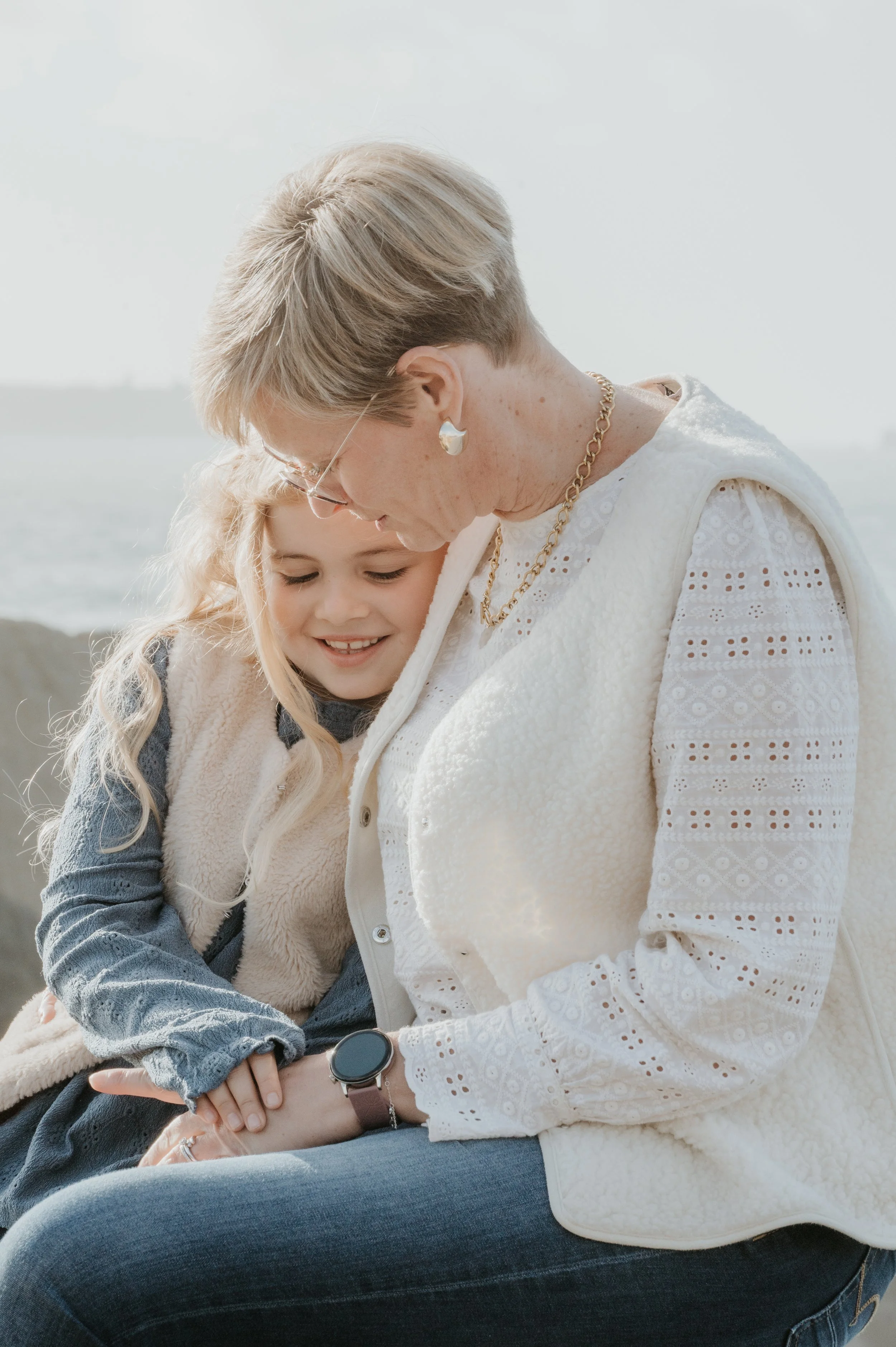 Une vieille femme et une jeune fille assises côte à côte, se regardant avec tendresse, près de la mer, par une journée ensoleillée.