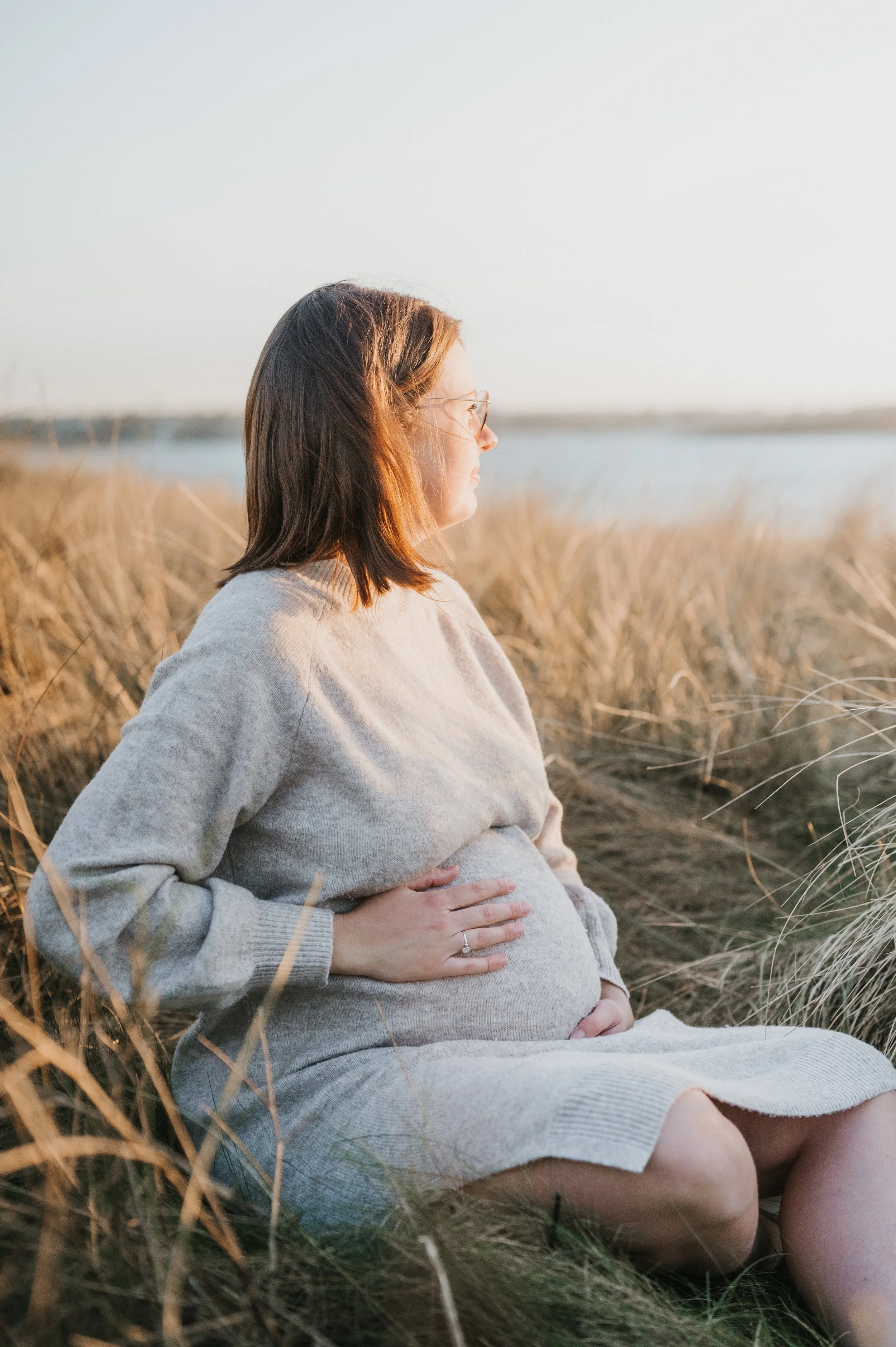 Femme enceinte assise dans un champ de herbes hautes, regardant vers le côté, avec un paysage d'eau en arrière-plan au coucher du soleil.