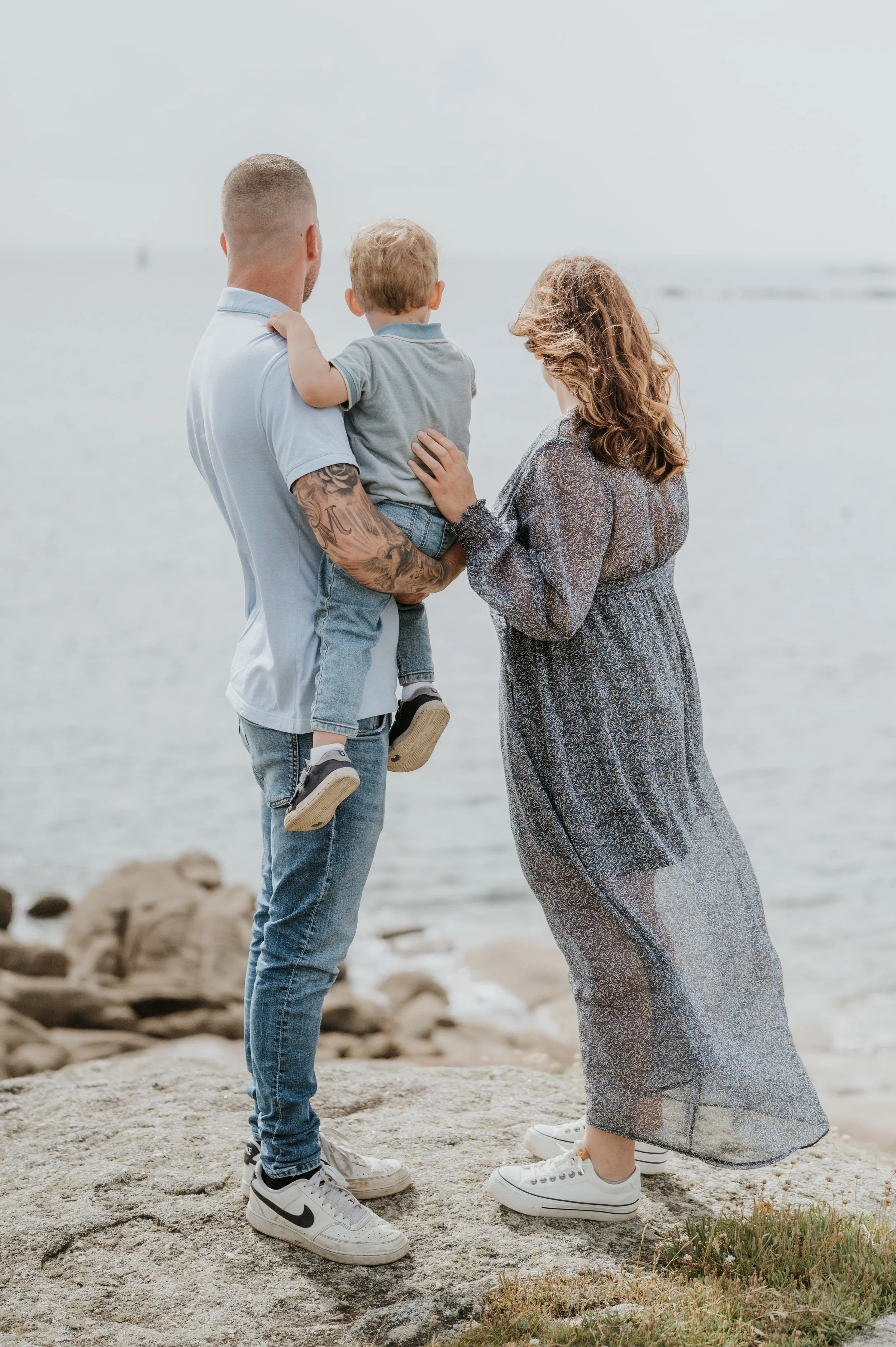 Une famille de trois personnes, un homme, une femme et un enfant, debout sur la plage près de l'océan, regardant à l'horizon. L'homme tient l'enfant dans ses bras, et la femme touche légèrement l'enfant avec une main, tous deux portant des vêtements 