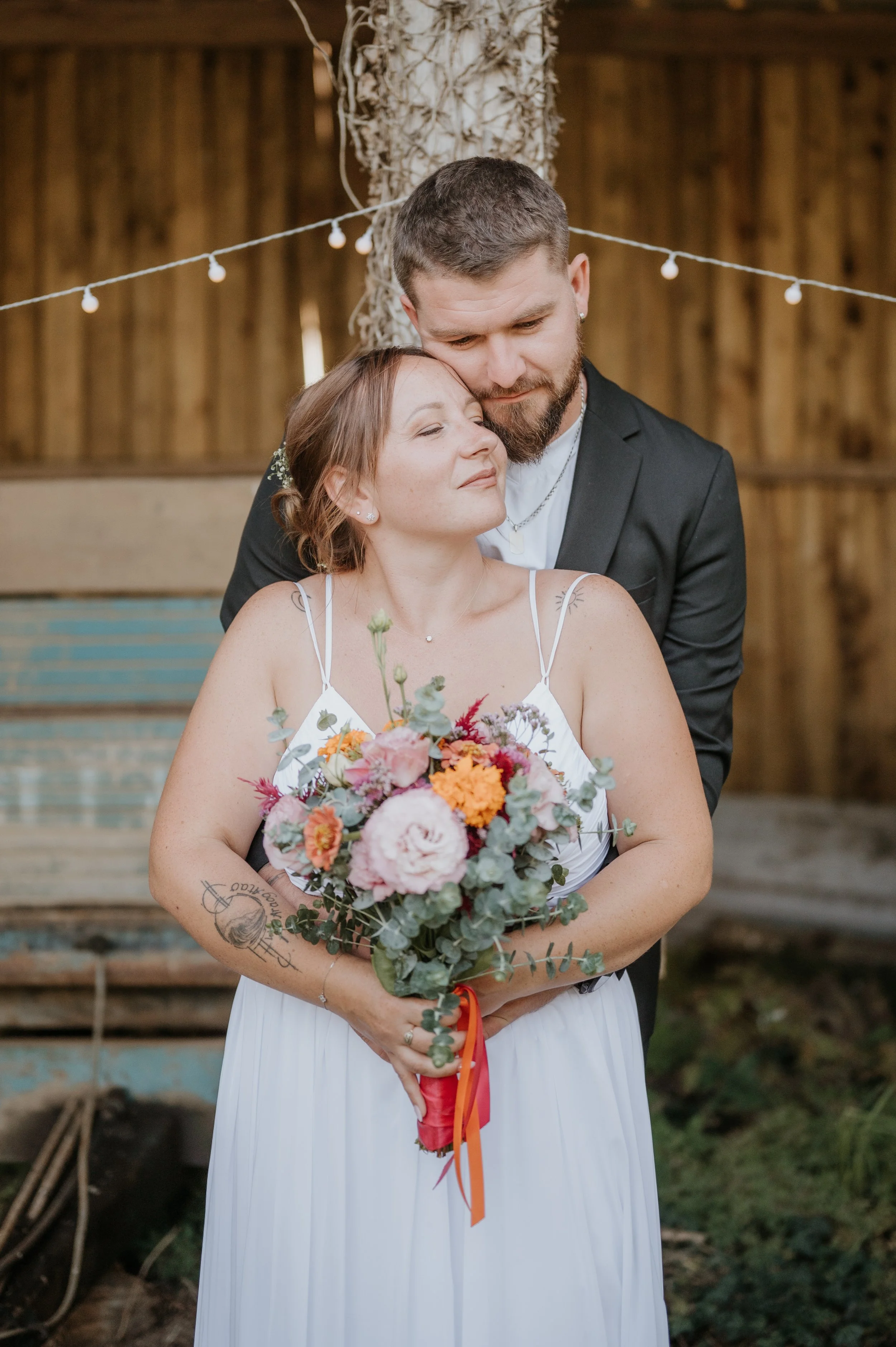 Un couple lors d'une cérémonie, la femme tient un bouquet de fleurs, et l'homme la serre dans ses bras, dans un cadre rustique en bois avec des guirlandes lumineuses.