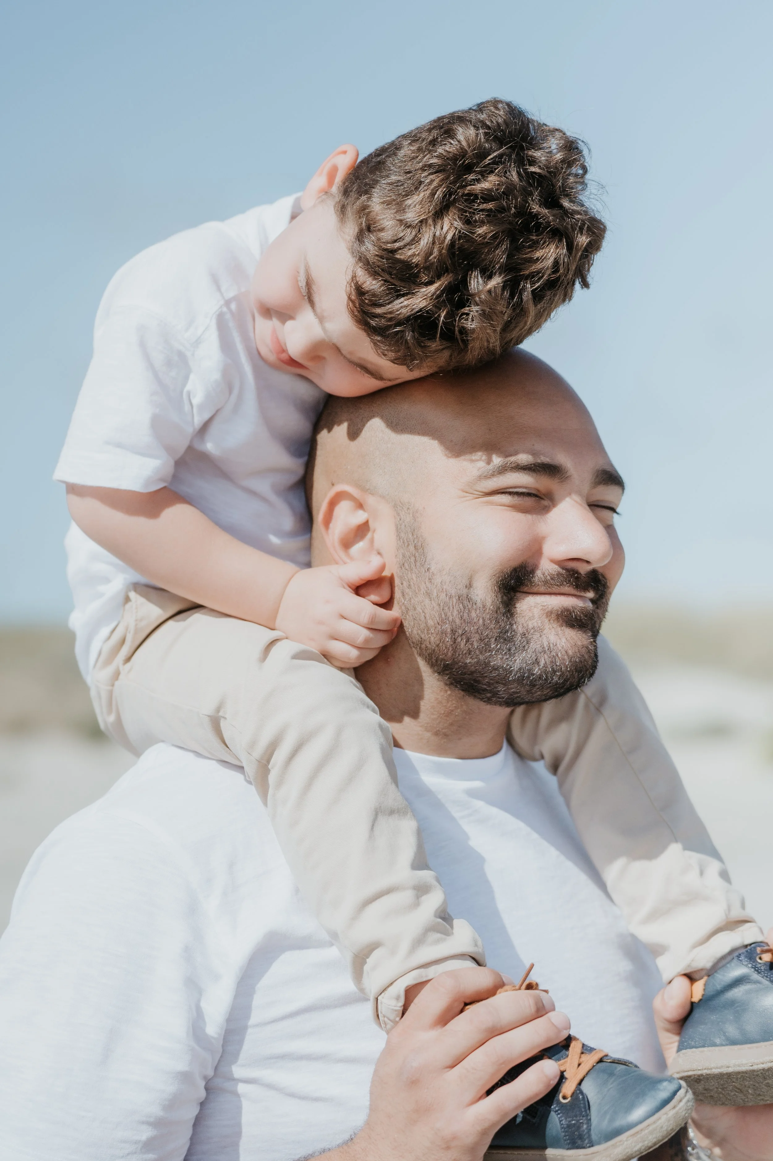 Un enfant positionné sur les épaules d'un homme, les yeux fermés et un sourire confortable, dans un environnement en extérieur, sous un ciel bleu clair.