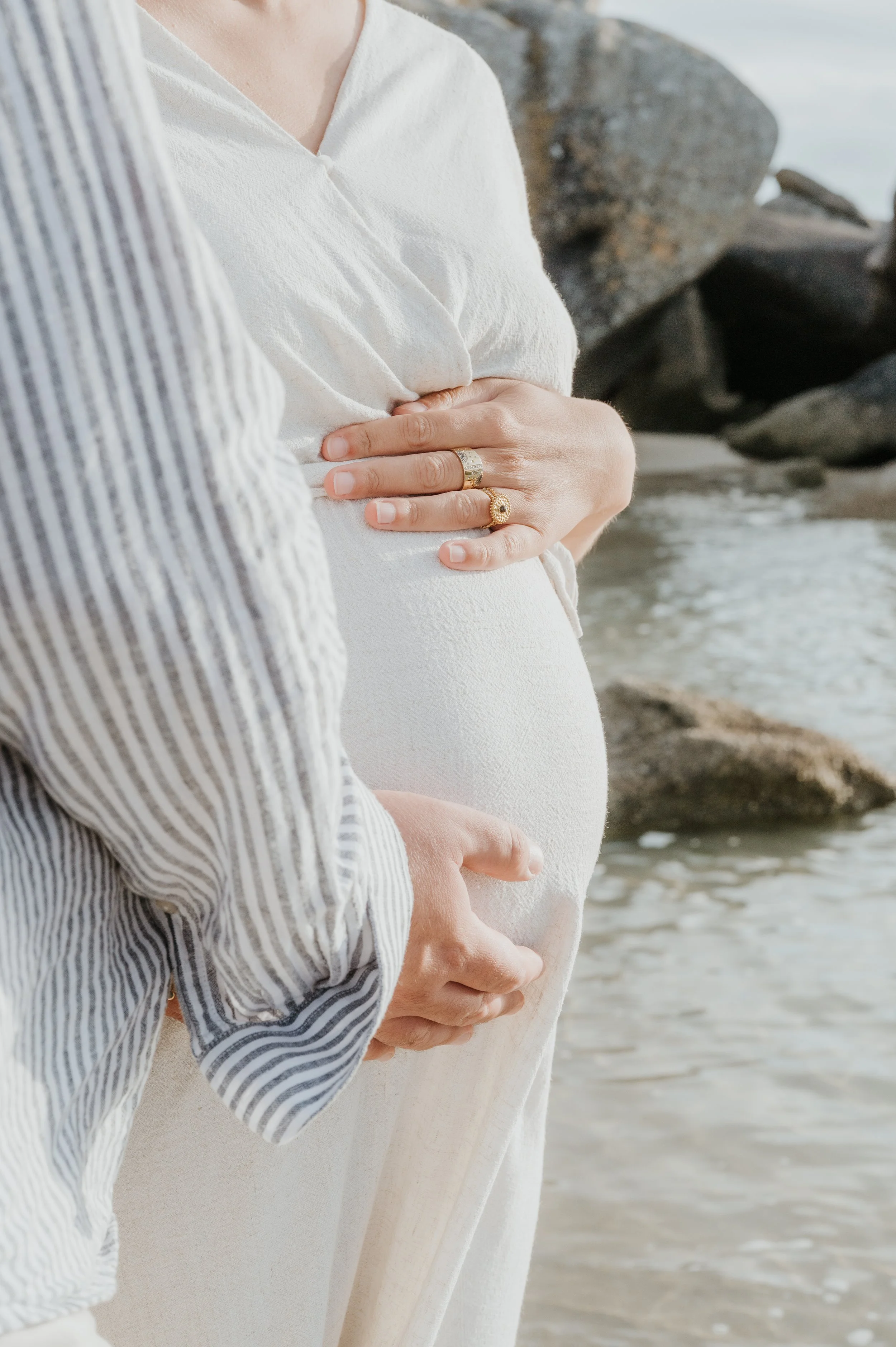 Deux personnes tenant le ventre d'une femme enceinte sur la plage, avec des rochers en arrière-plan.