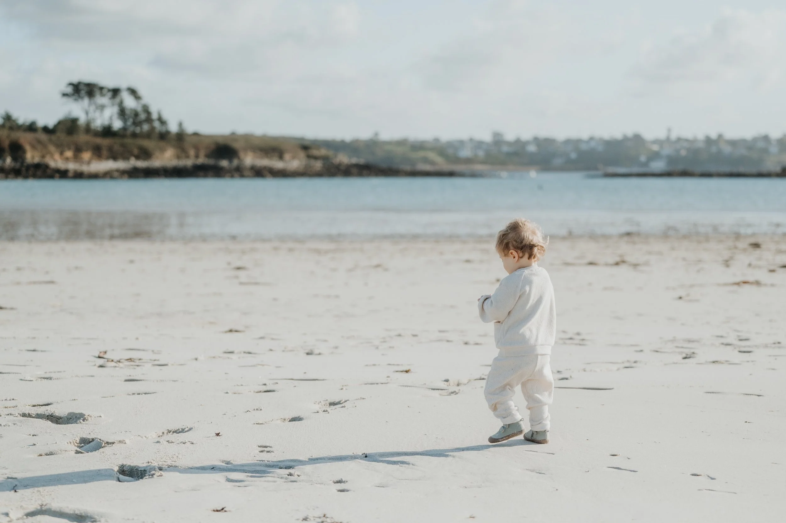 Un enfant marchant sur la plage de sable, portant des vêtements légers, face à la mer avec un ciel nuageux.