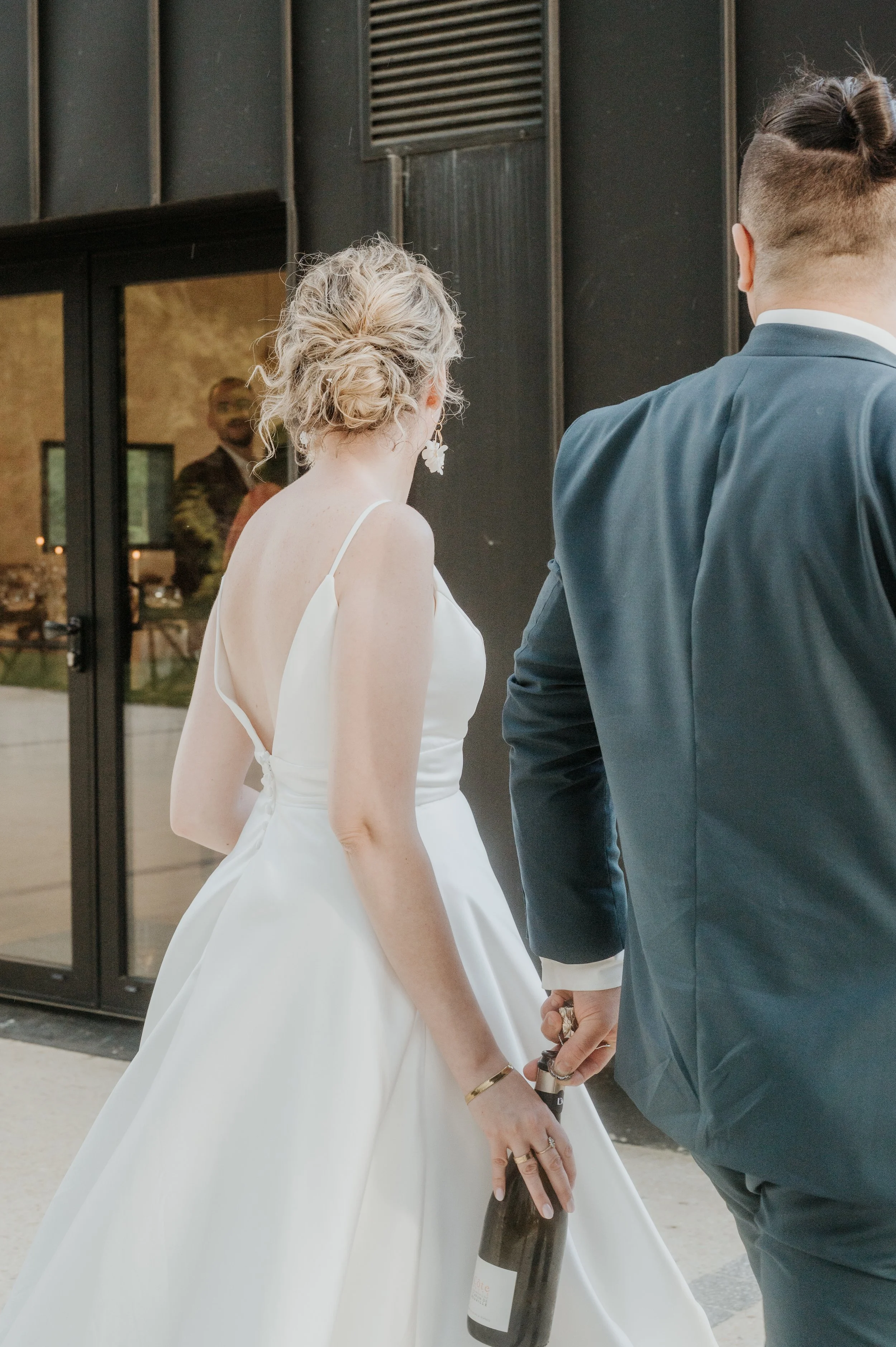 Un couple lors d'un mariage, la femme en robe blanche et l'homme en costume bleu, se tenant par la main. La femme porte des boucles d'oreilles en forme de fleurs et un bracelet doré.