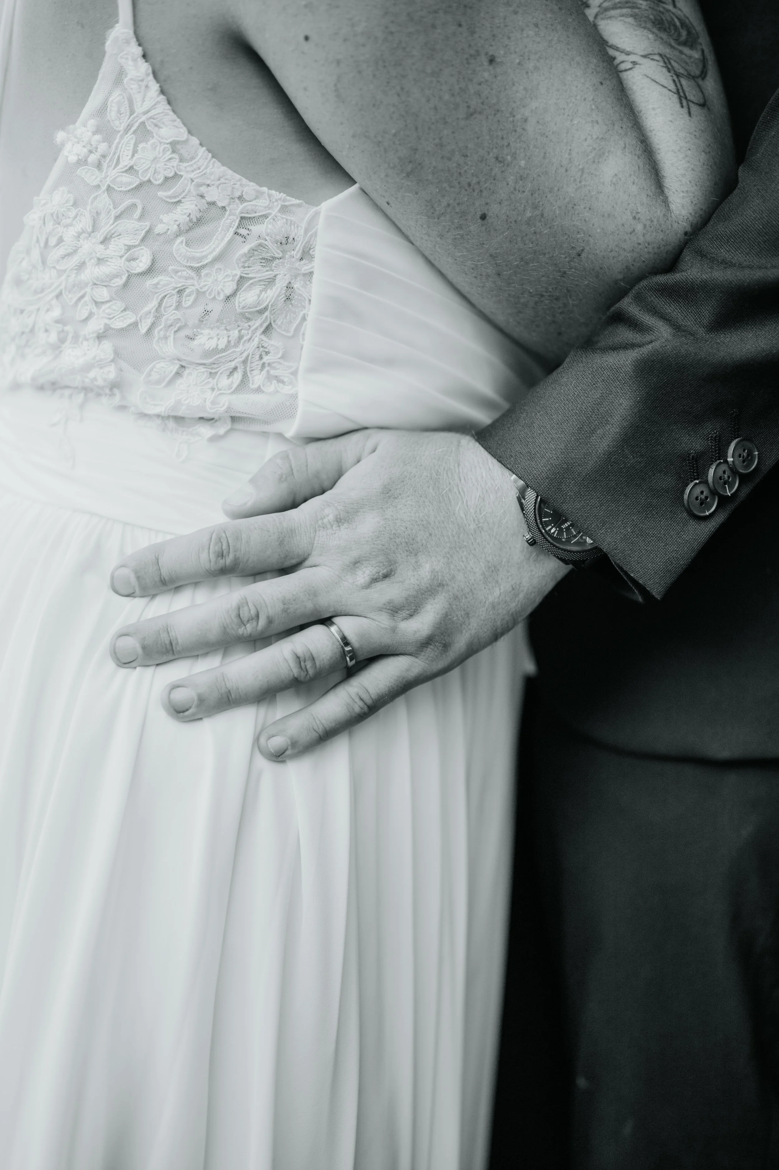 Gros plan d'un mariage, montrant la main d'un homme portant une alliance, posé sur la robe blanche d'une femme avec des détails en dentelle et tissu plissé, lors d'une cérémonie nuptiale.