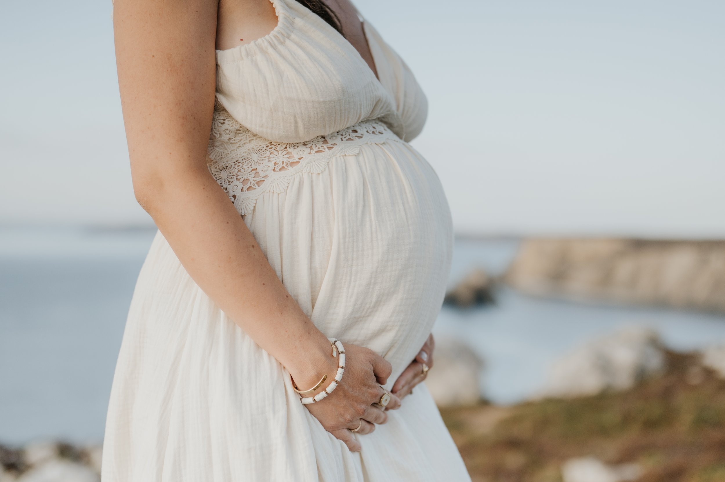 Une femme enceinte portant une robe blanche avec des détails en dentelle, tenant ses mains sur son ventre, sur une plage au bord de l'océan.