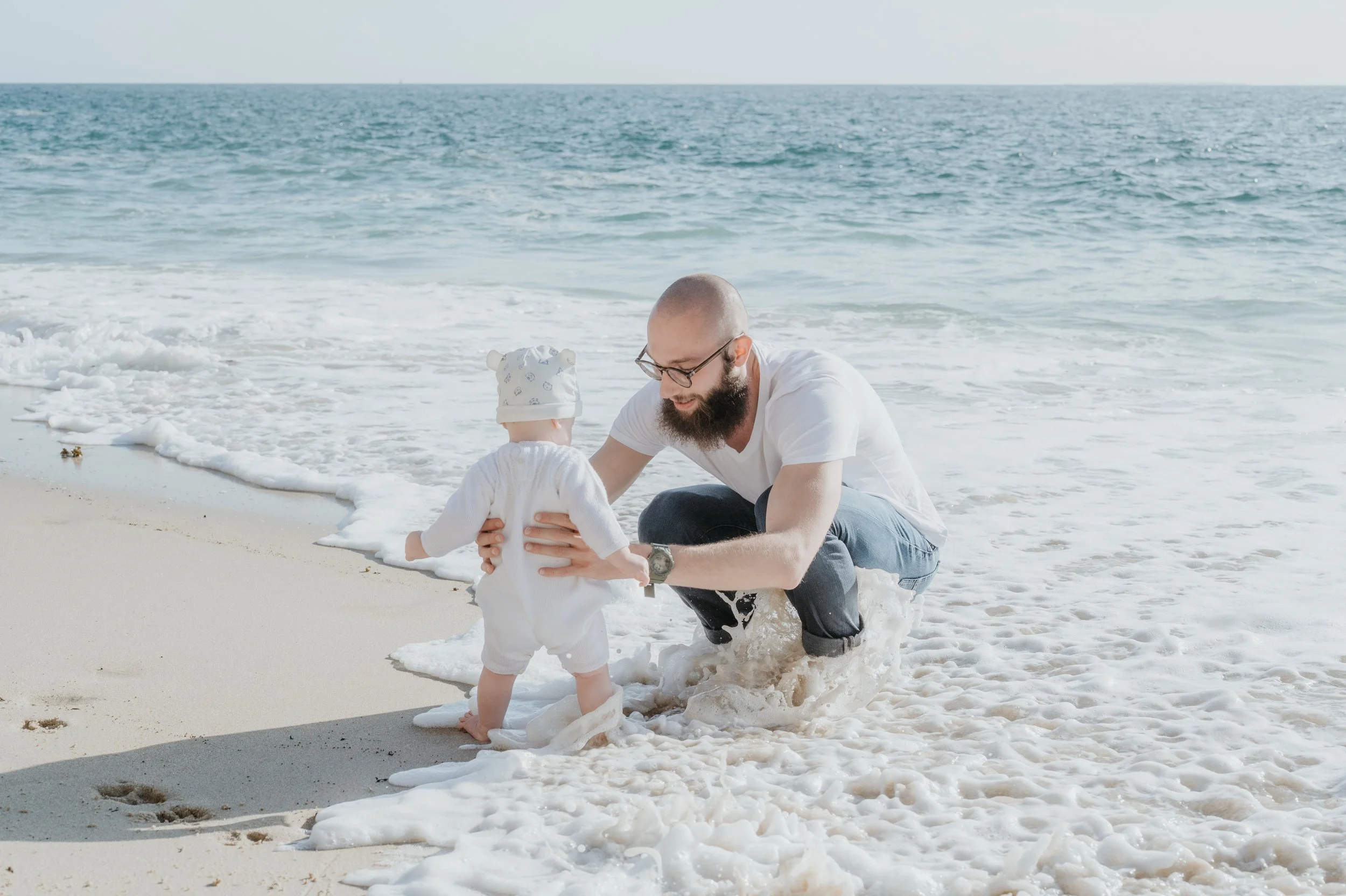 Un homme avec une barbe et des lunettes joue avec un bébé sur une plage, tous deux habillés en blanc, près de l'eau et du sable.