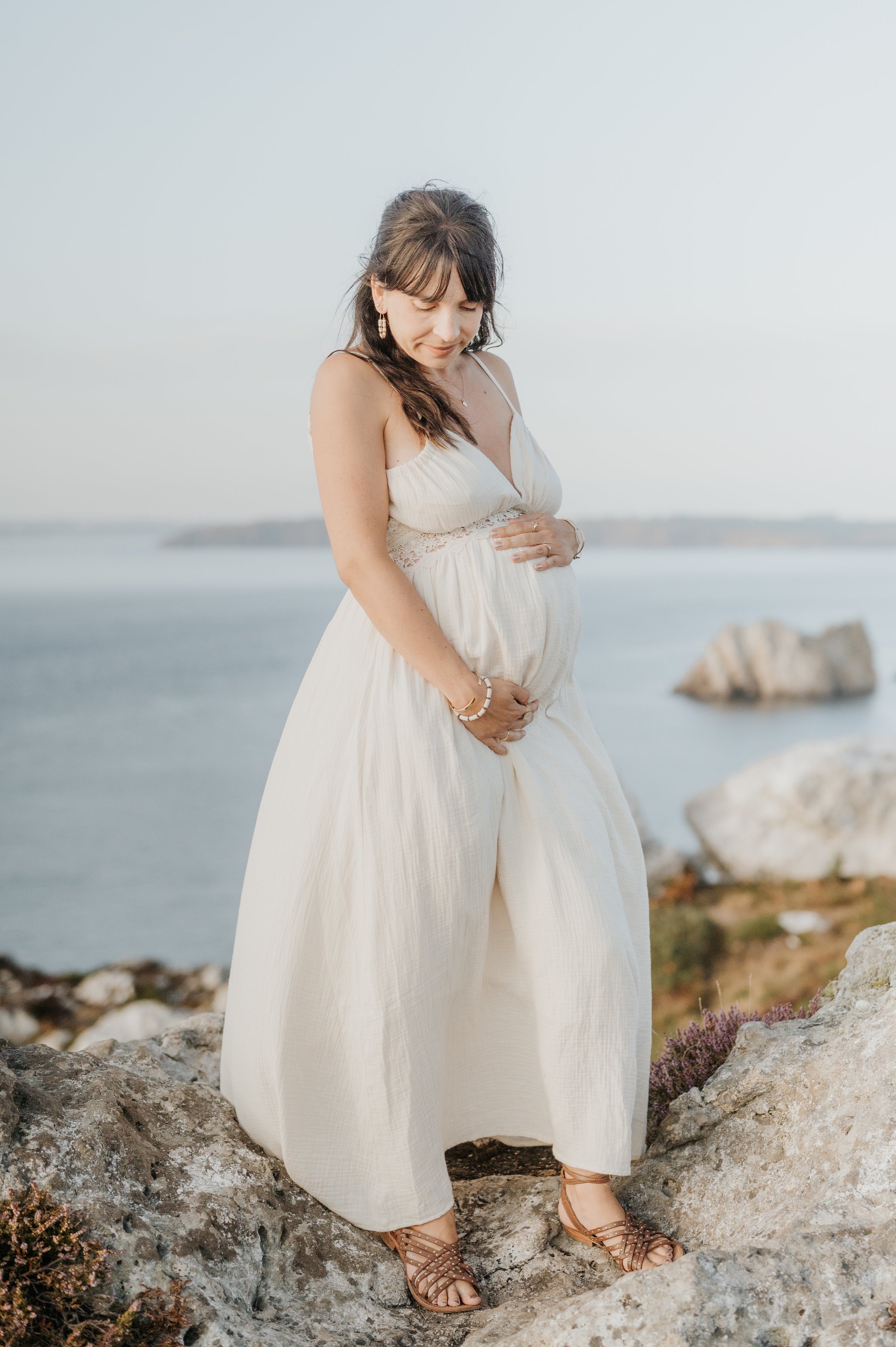 Une femme enceinte portant une robe blanche longue, coiffée de cheveux bruns, se tient sur des rochers près de la mer, regardant vers le bas avec les yeux fermés.