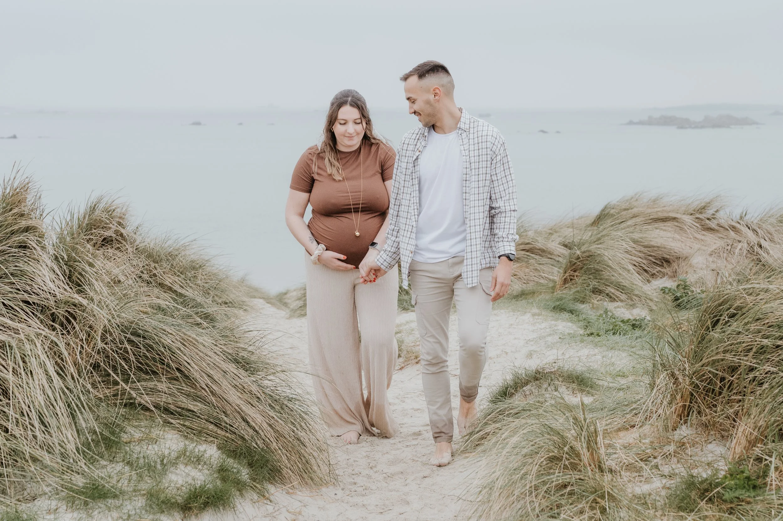 Un couple marchant main dans la main sur une plage avec des herbes hautes, la femme est enceinte et le paysage est brumeux avec la mer en arrière-plan.