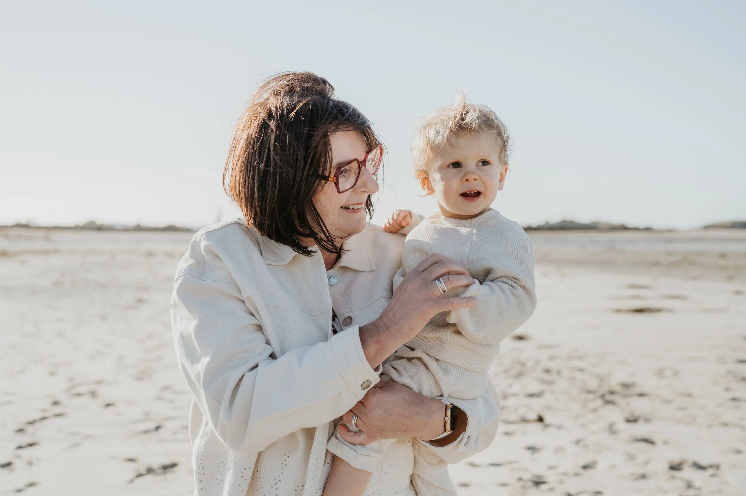 Une femme souriante tenant un jeune enfant blond dans ses bras sur une plage ensoleillée.