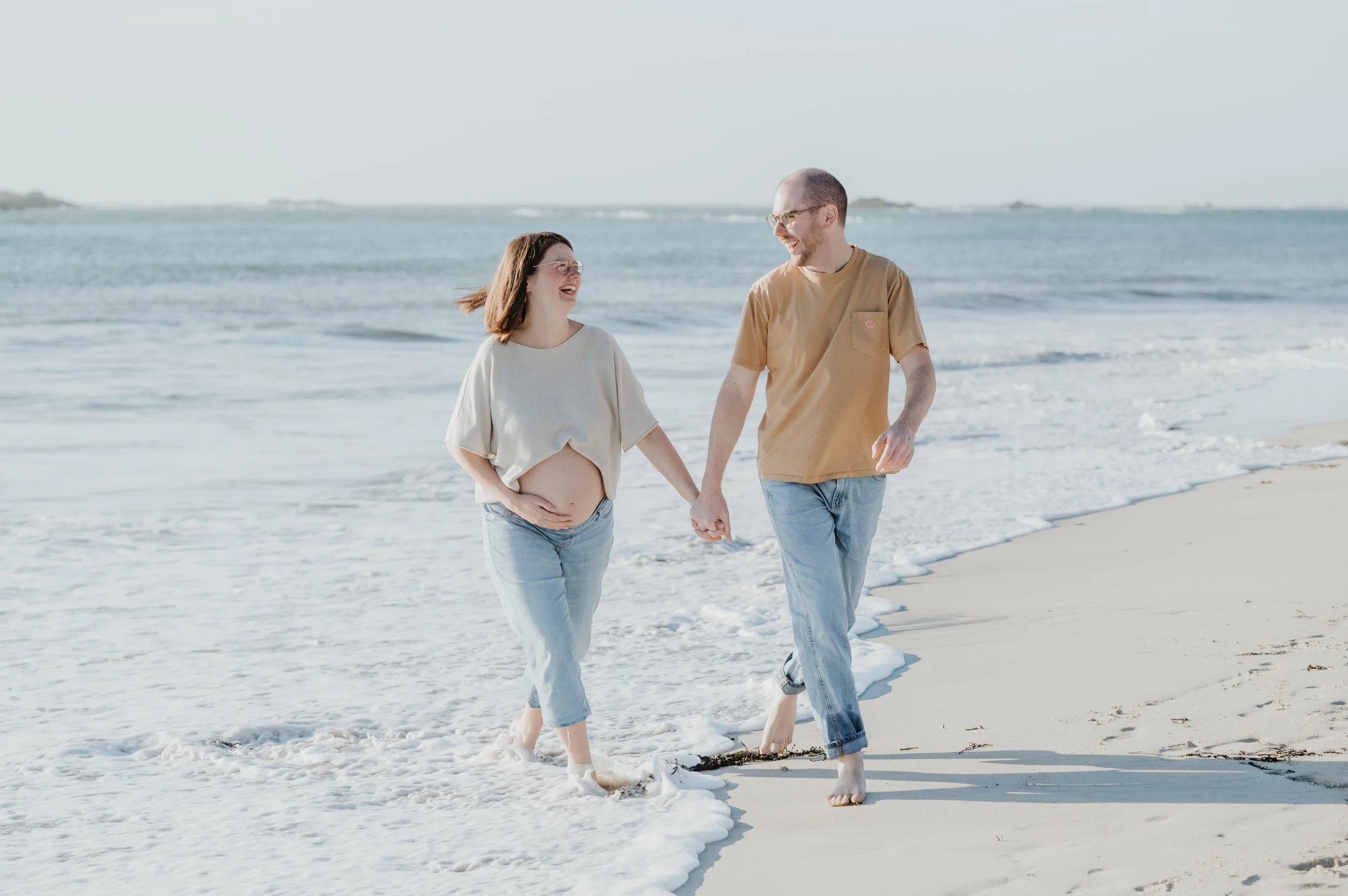 Un couple marche main dans la main le long de la plage, avec la mer en arrière-plan, sous un ciel clair.