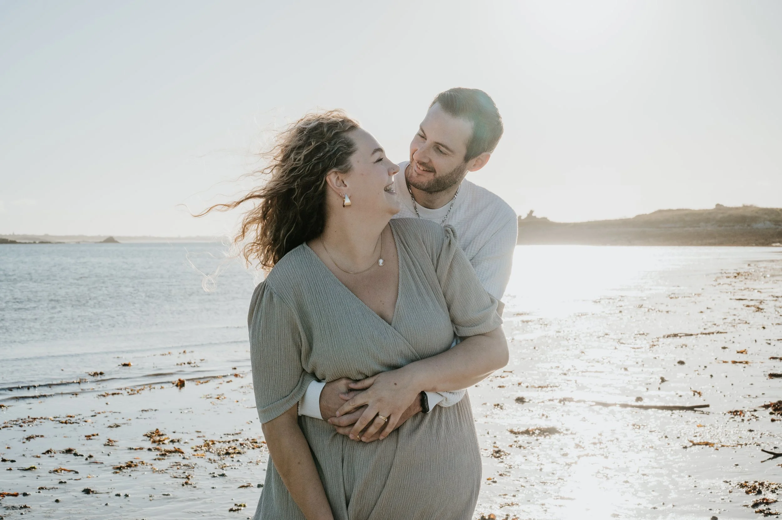 Un couple souriant se tenant dans les bras sur la plage au coucher du soleil, avec la mer en arrière-plan.