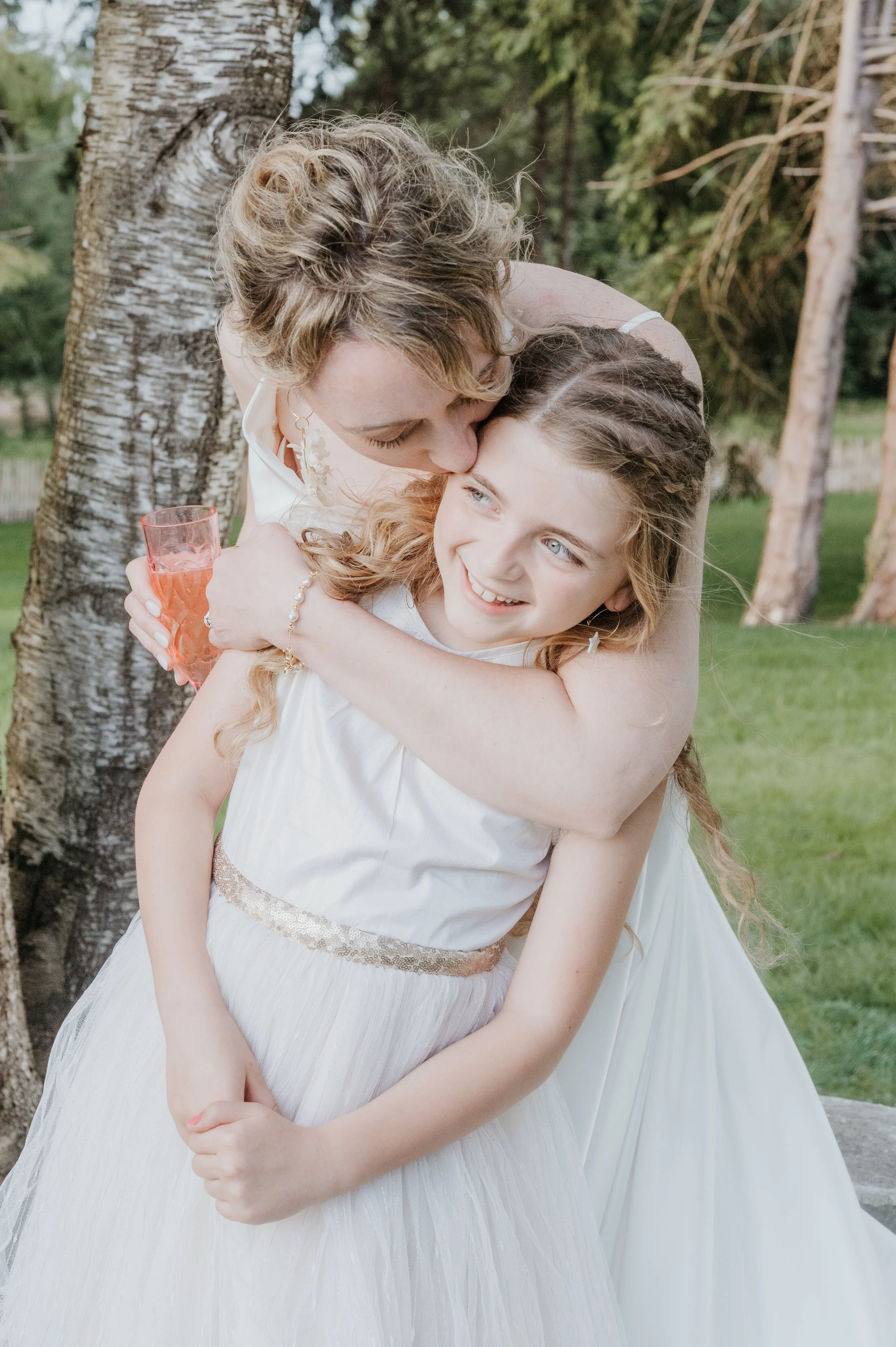 Une femme et une jeune fille s'embrassent dans un parc, toutes deux portant des robes blanches, entourées d'arbres.