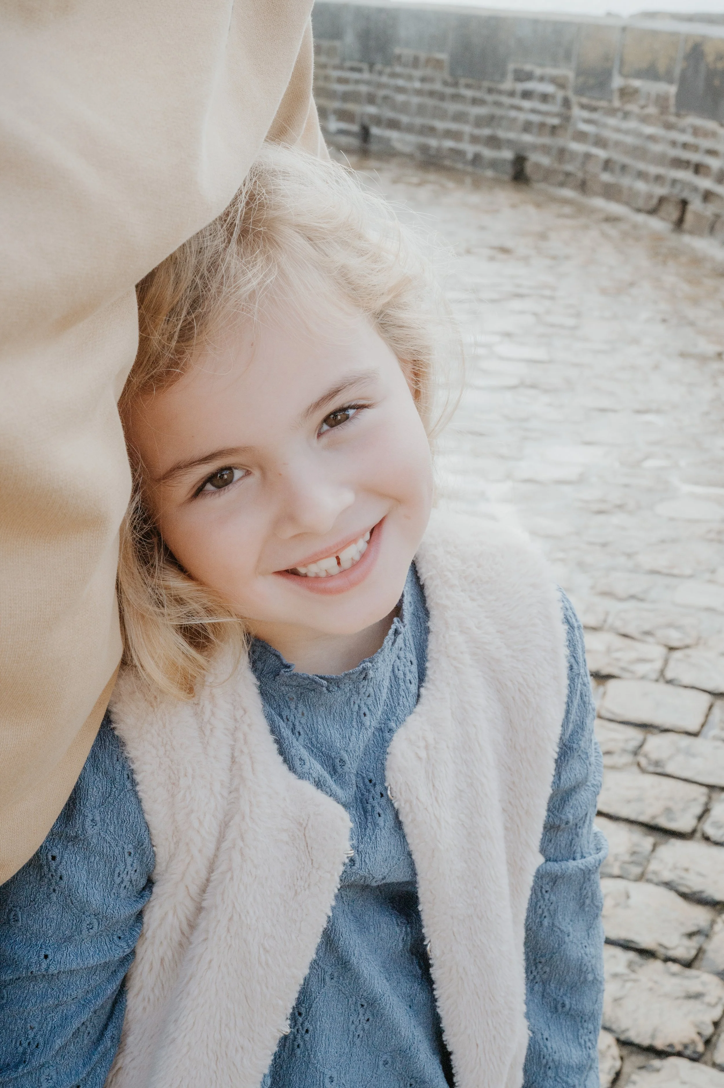 Une jeune fille souriante avec des cheveux blonds, portant une veste en jean et un gilet en peluche, se tient près d'une muraille en pierre, dans un environnement extérieur.
