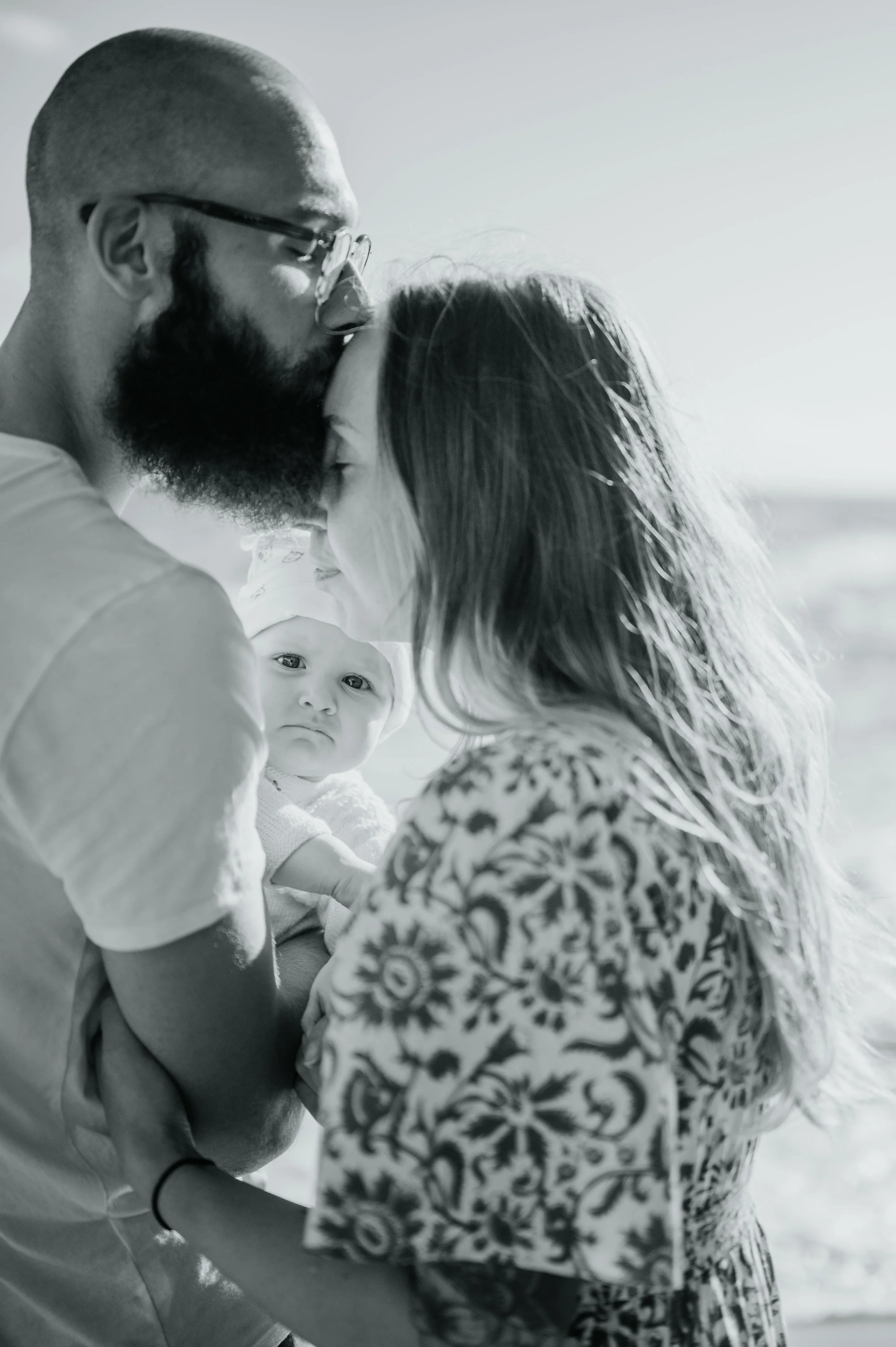 Un couple embrasse tendrement avec un bébé regardant vers la caméra, en extérieur, en noir et blanc.