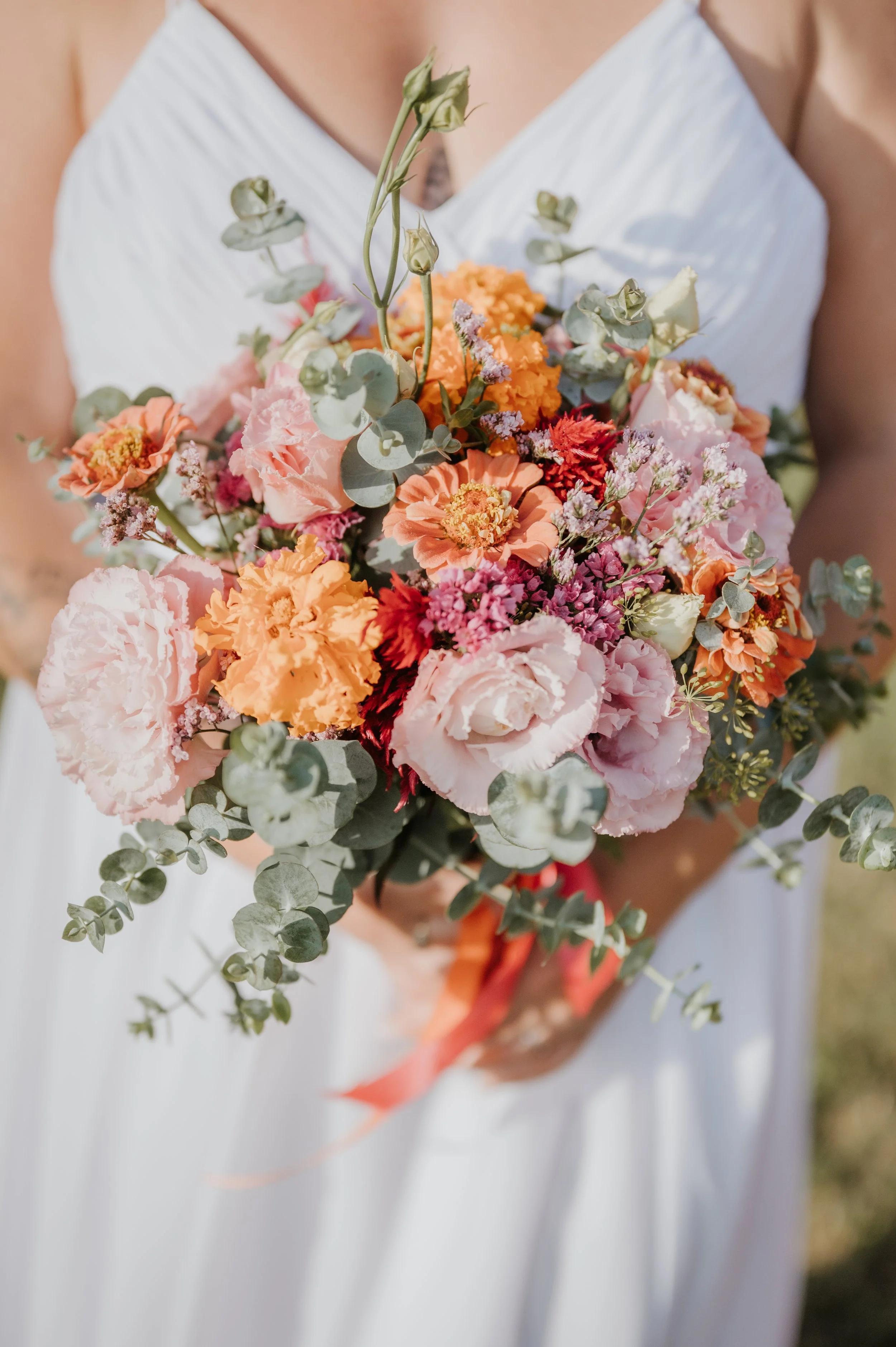Une personne en robe blanche tient un bouquet de fleurs de différentes couleurs, notamment rose, orange, blanc, rouge et violet, avec des feuilles de eucalyptus, lors d'une occasion spéciale, probablement un mariage.