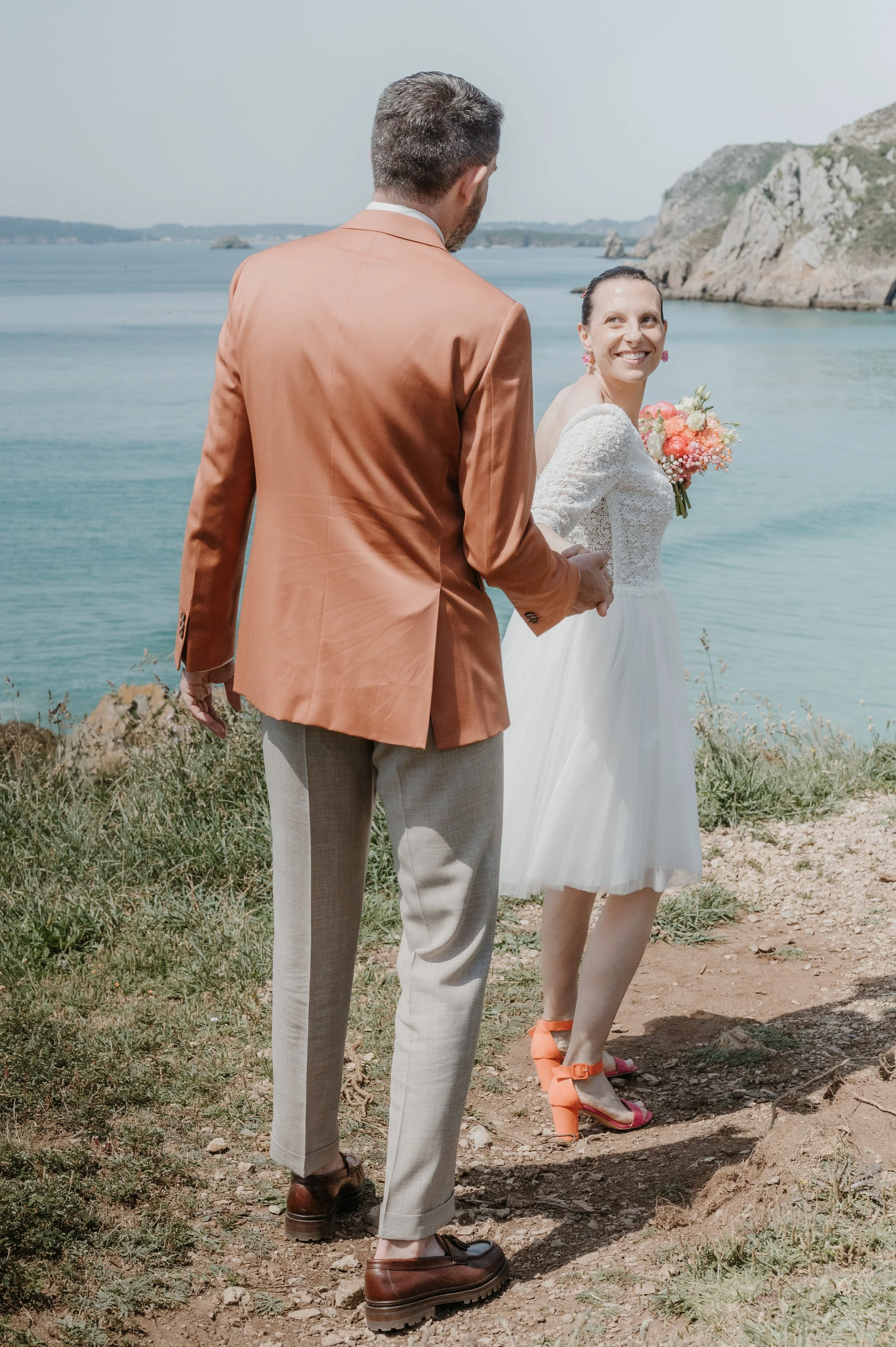 Un homme en costume beige et une femme en robe blanche de mariage en bord de mer, souriant, avec des rochers et la mer en arrière-plan.