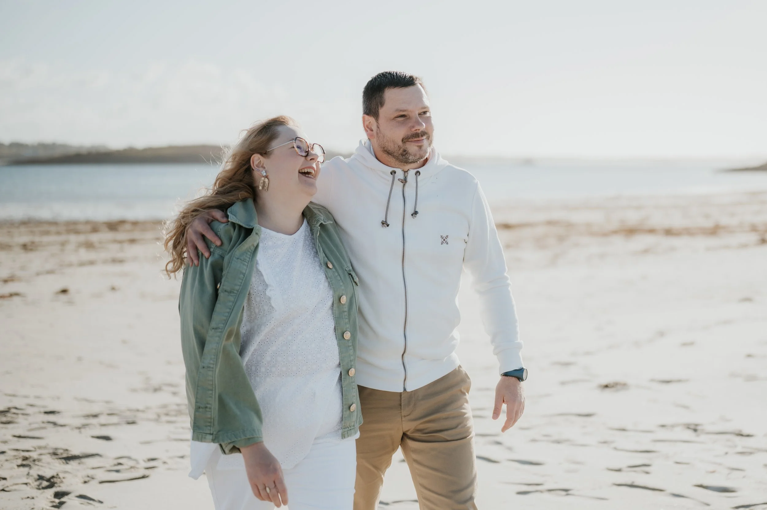 Un couple marche main dans la main sur la plage en souriant, en tenue décontractée, sous un ciel clair.