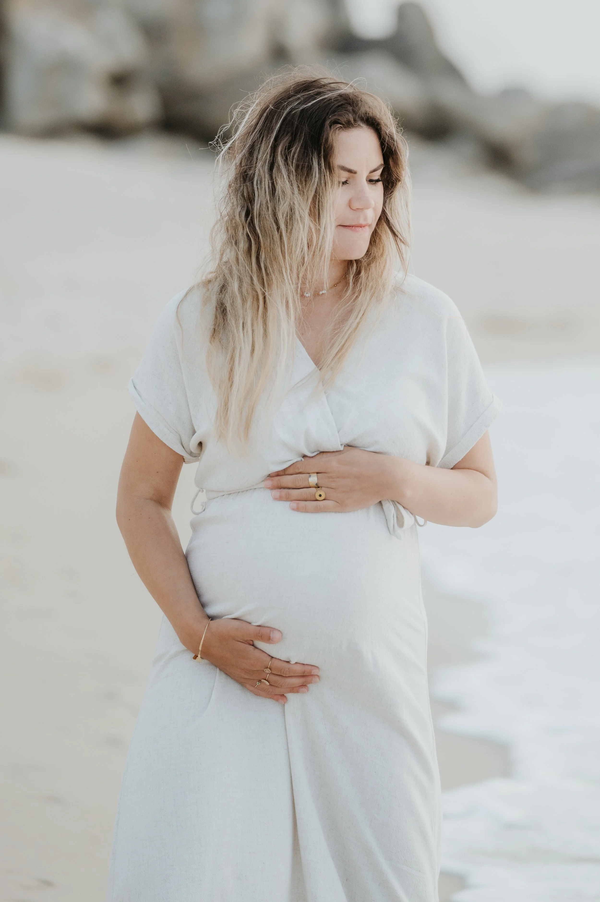 Une femme enceinte portant une robe blanche, se tenant la main sur le ventre, se trouve sur une plage avec des rochers en arrière-plan.