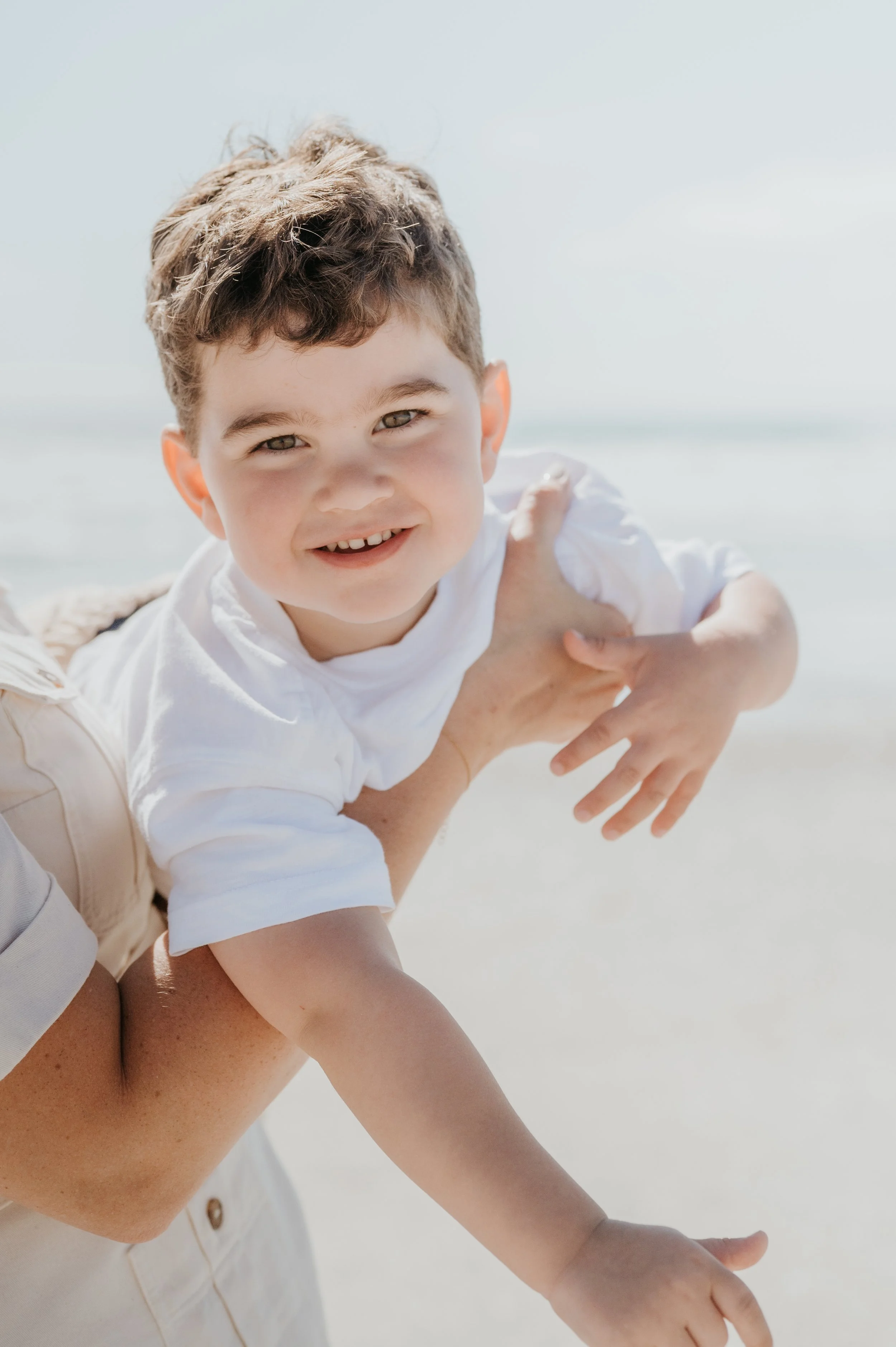 Un enfant souriant avec les cheveux bruns, portant un t-shirt blanc, joue sur la plage sous un ciel ensoleillé.