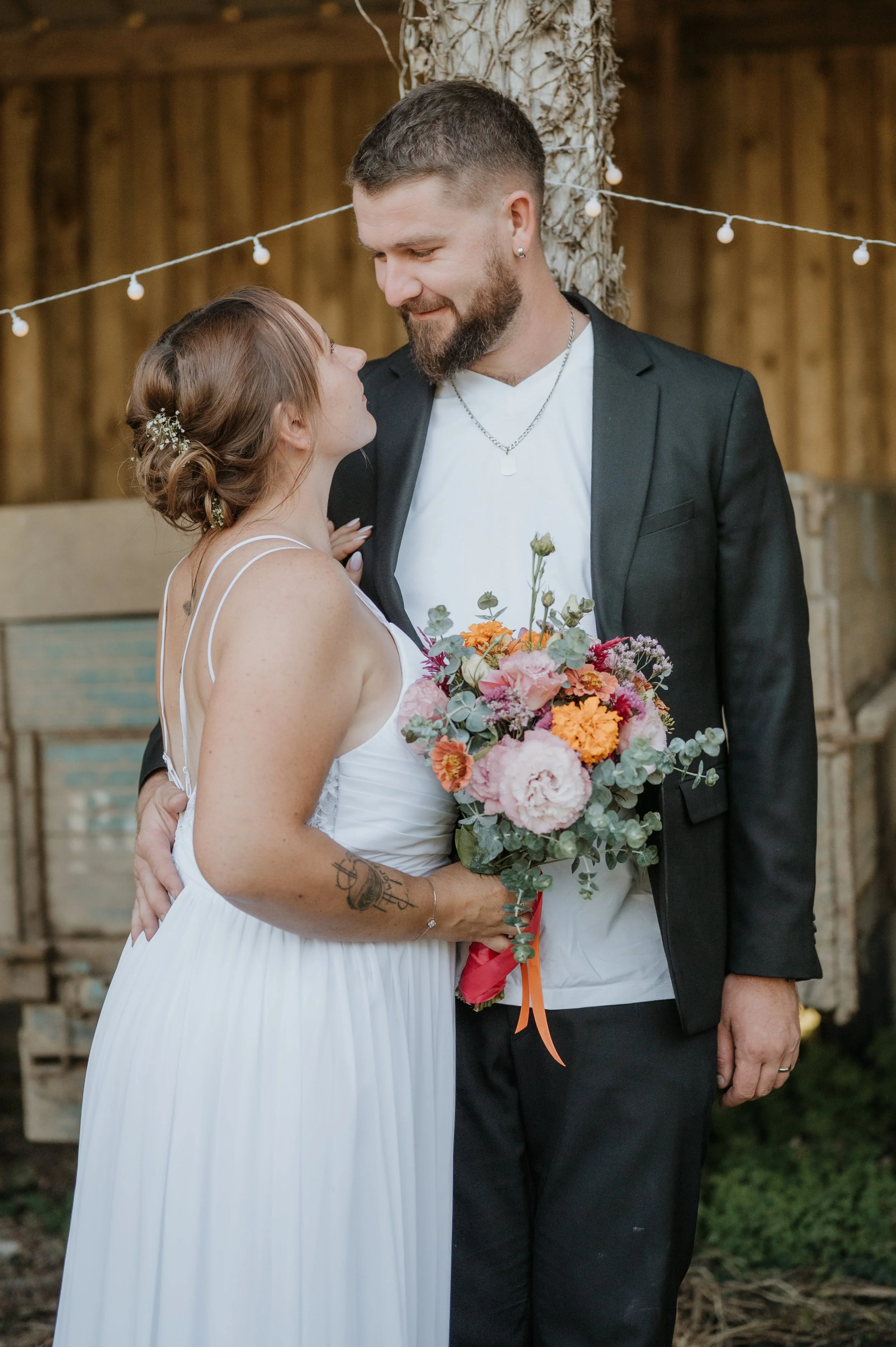 Un couple de jeunes mariés, la femme en robe blanche et le homme en costume noir, se regardant tendrement lors d'une cérémonie en extérieur, avec un bouquet de fleurs colorées.