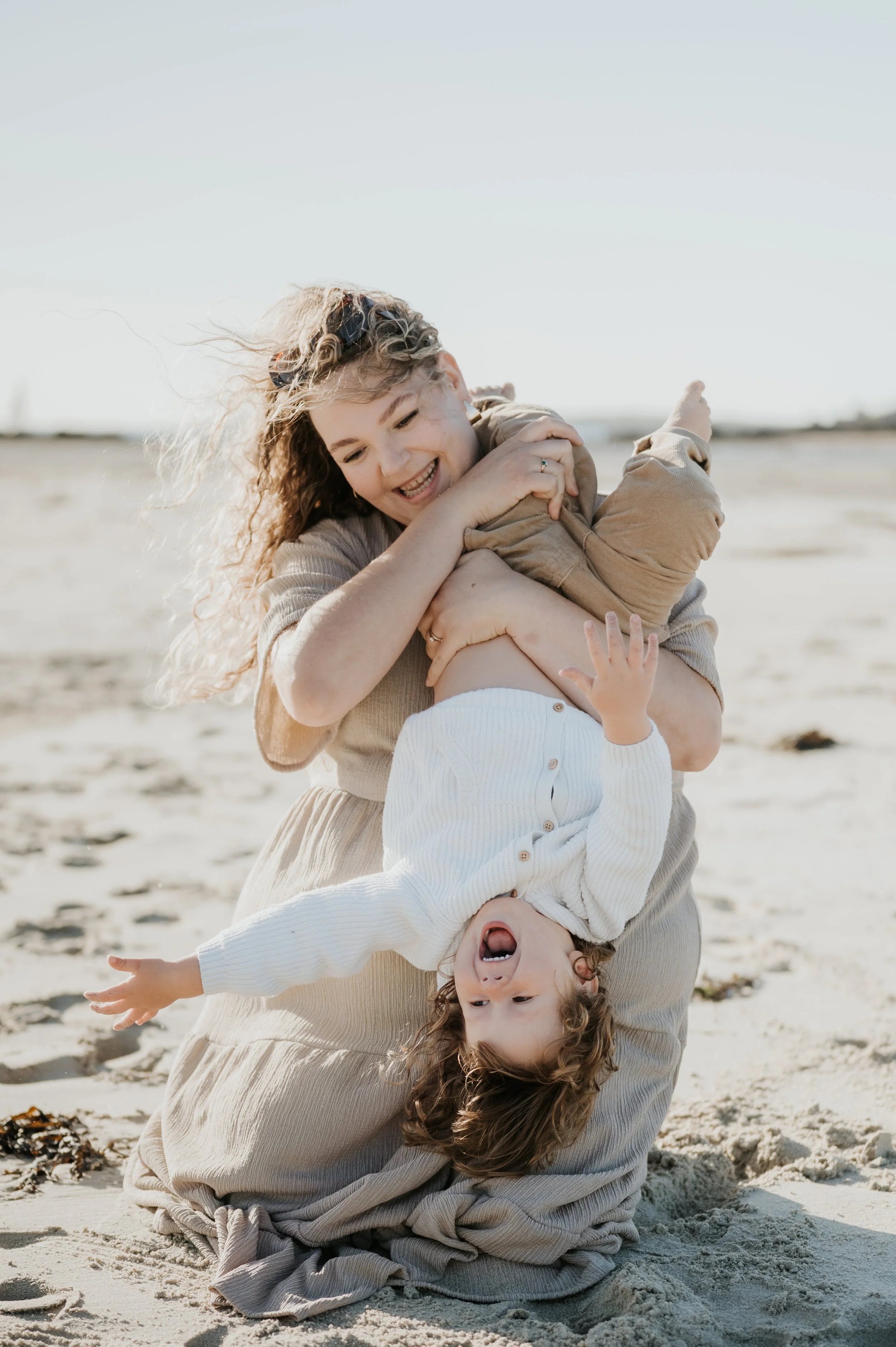 Une femme et deux enfants jouent sur la plage, la femme tient un enfant en l'air et l'autre enfant est à l'envers, souriant et riant dans le sable.