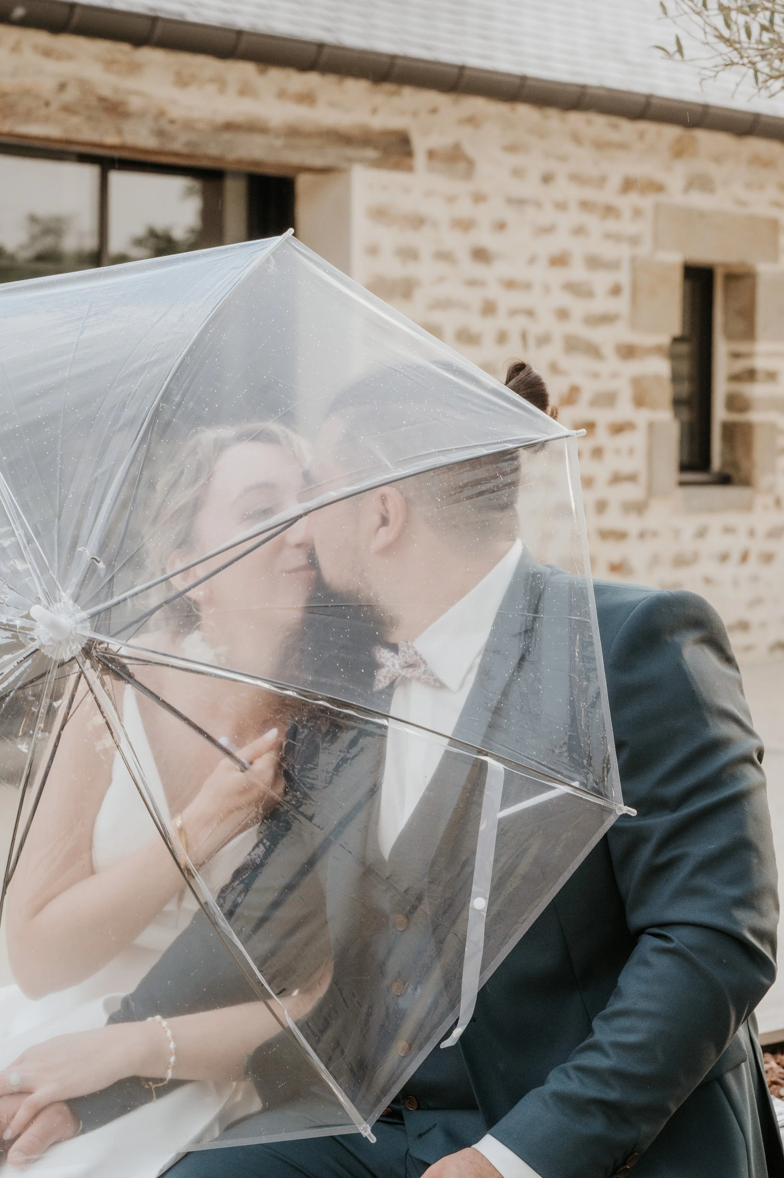 Un couple en habits formels partage un baiser sous un parapluie transparent, en bordure d'une maison en pierre.